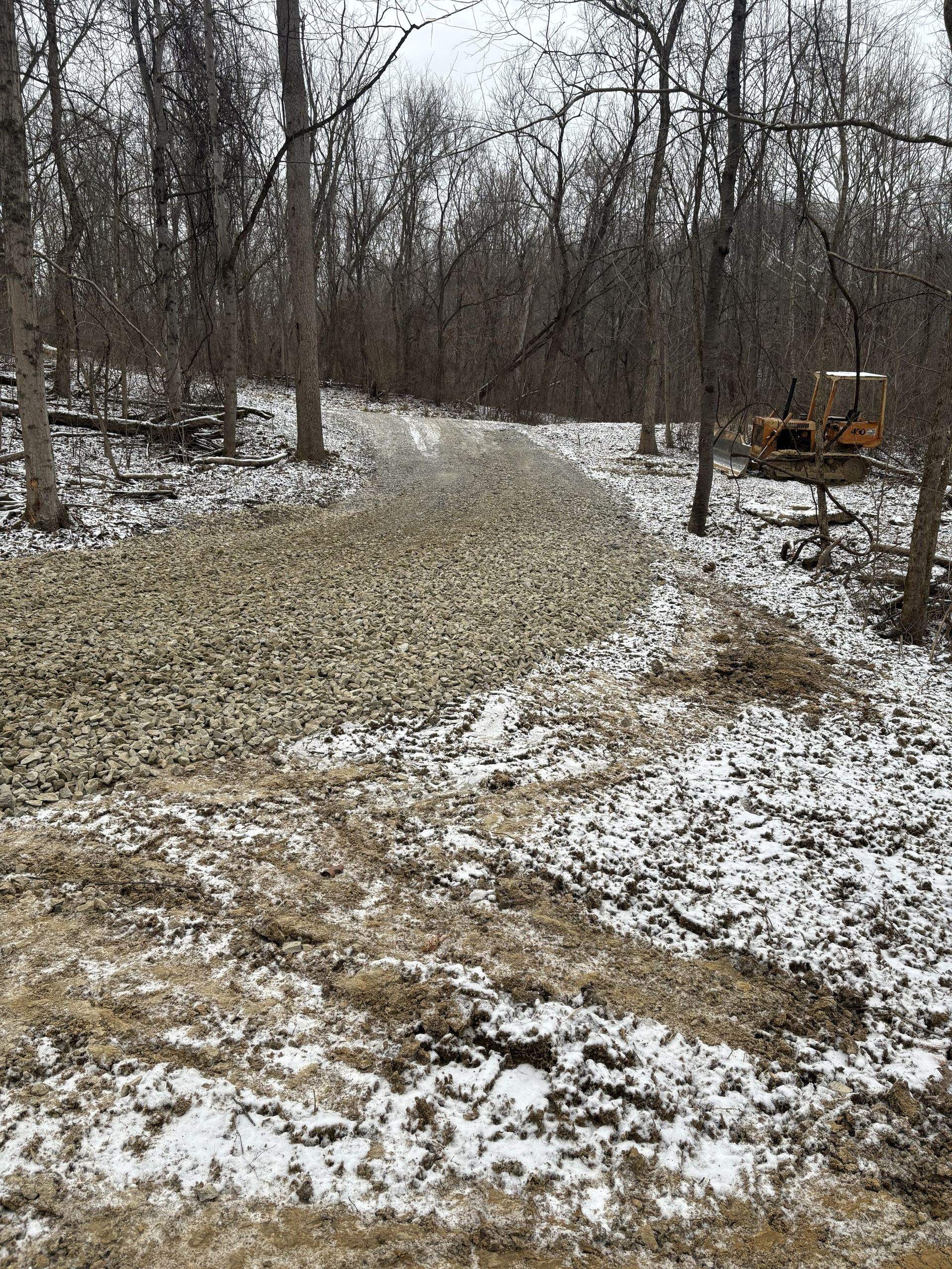 Snow-dusted wooded trail curving through bare trees, with an orange utility vehicle at right.
