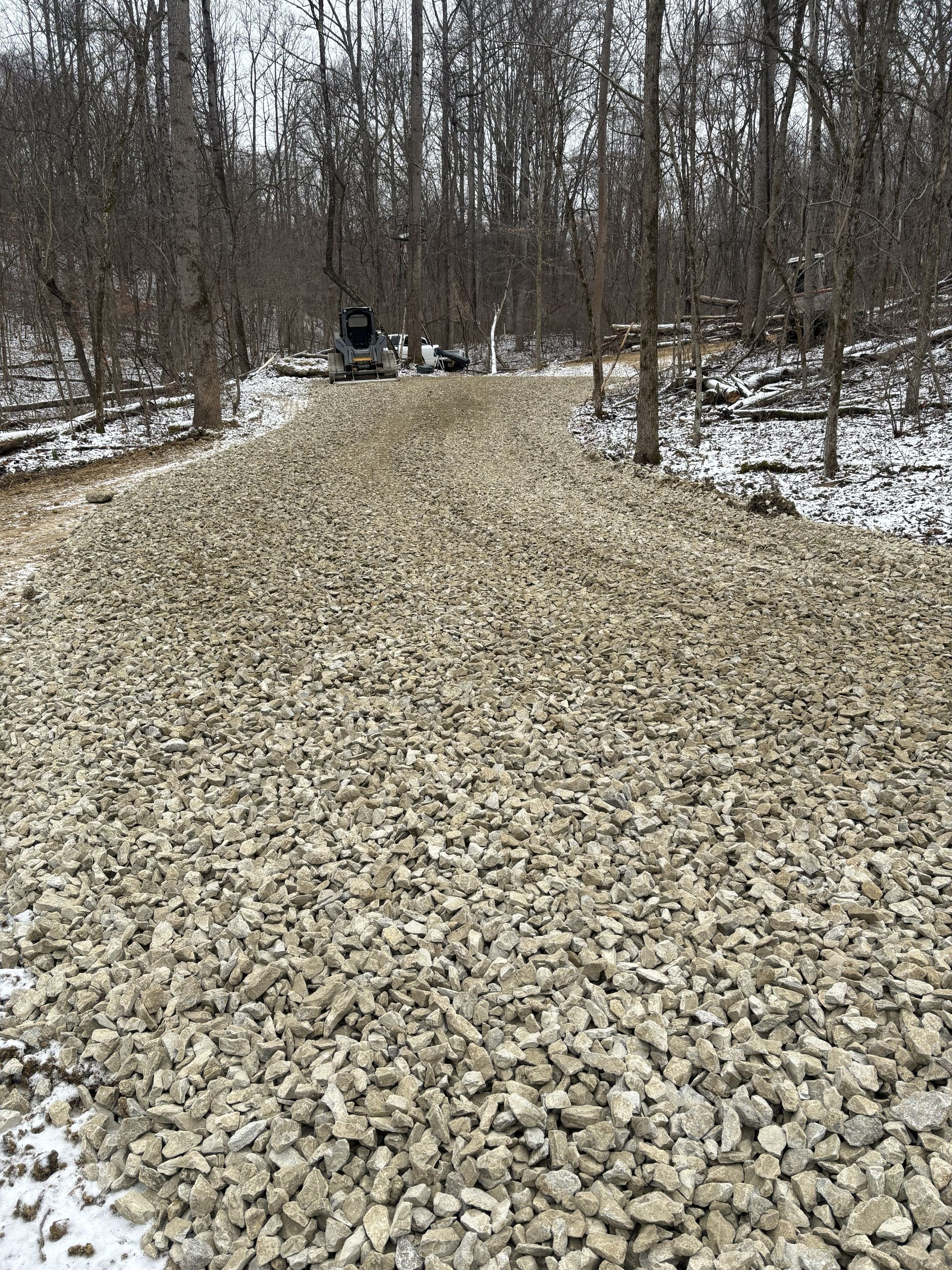 Gravel road through a leafless wooded area with light snow on the ground and a vehicle in the distance
