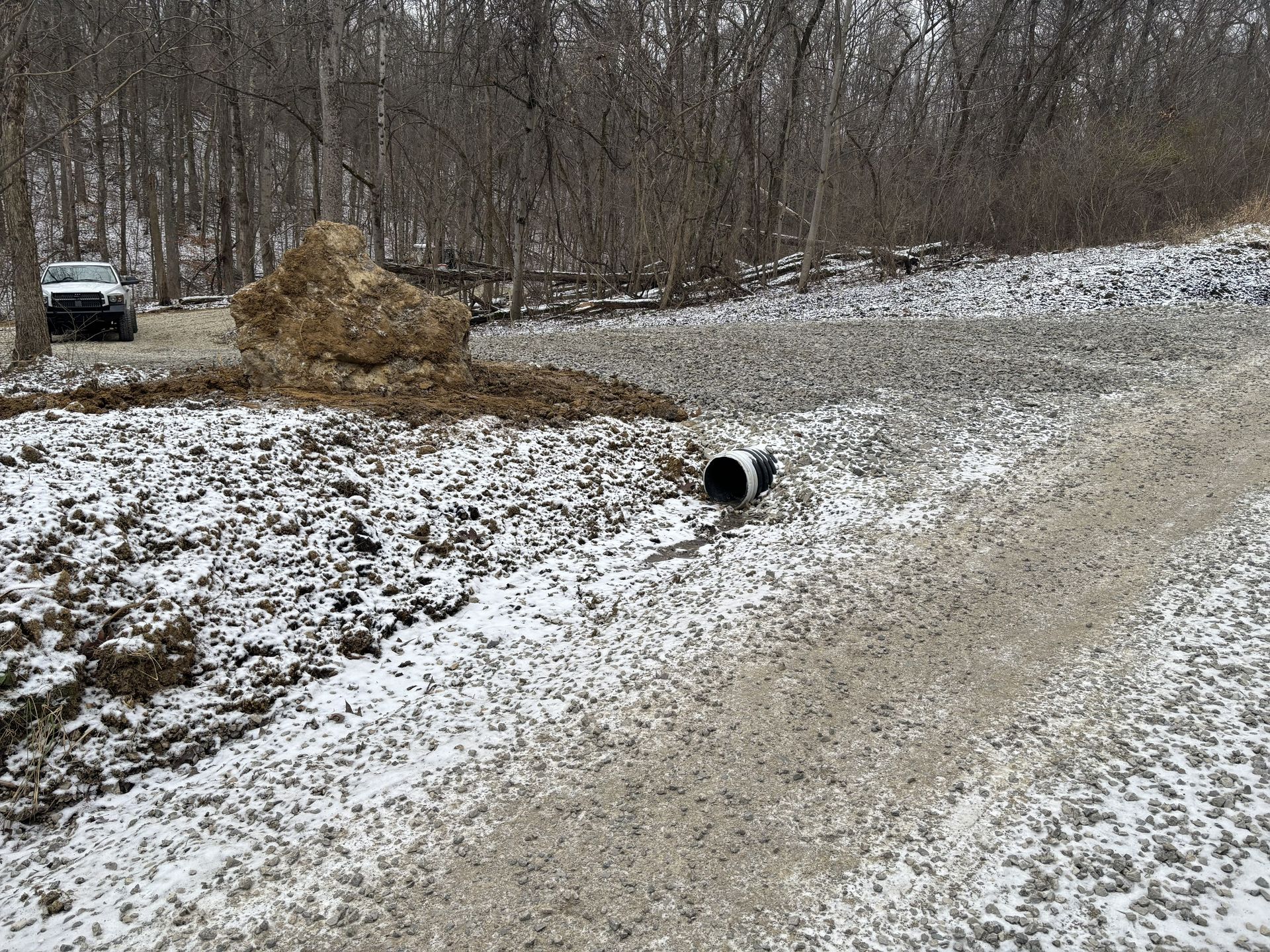 Snow-dusted rural road beside a wooded embankment with a drainage pipe and dirt pile