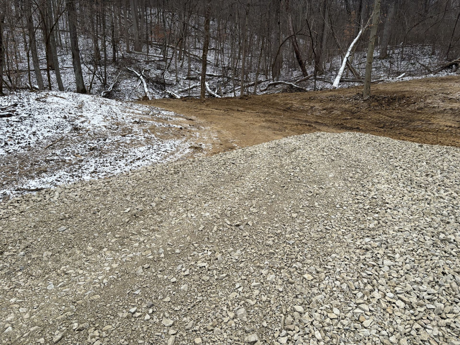 Gravel road with muddy brown patch beside a snow-dusted wooded hillside