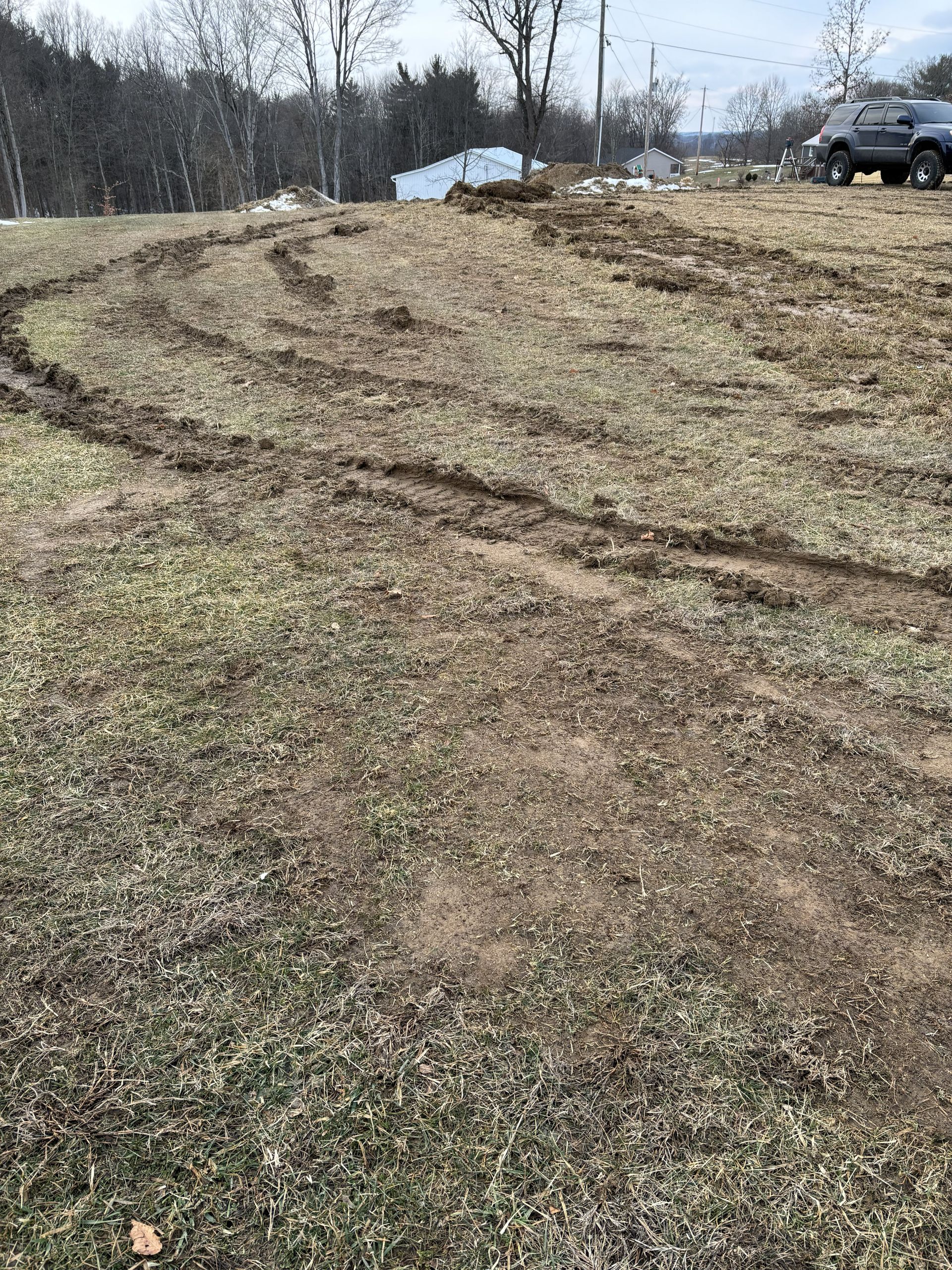 Rutted dirt and grass field with tire tracks leading toward trees and a parked truck on a cloudy day