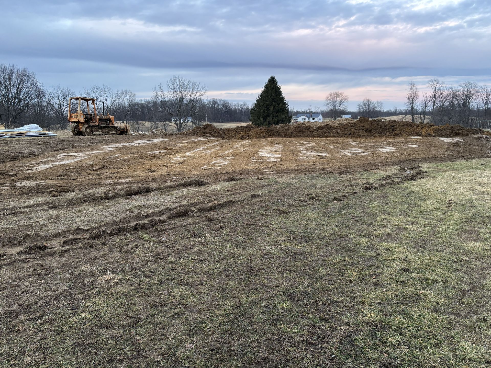 Bulldozer grading a muddy field under a cloudy sky at dusk