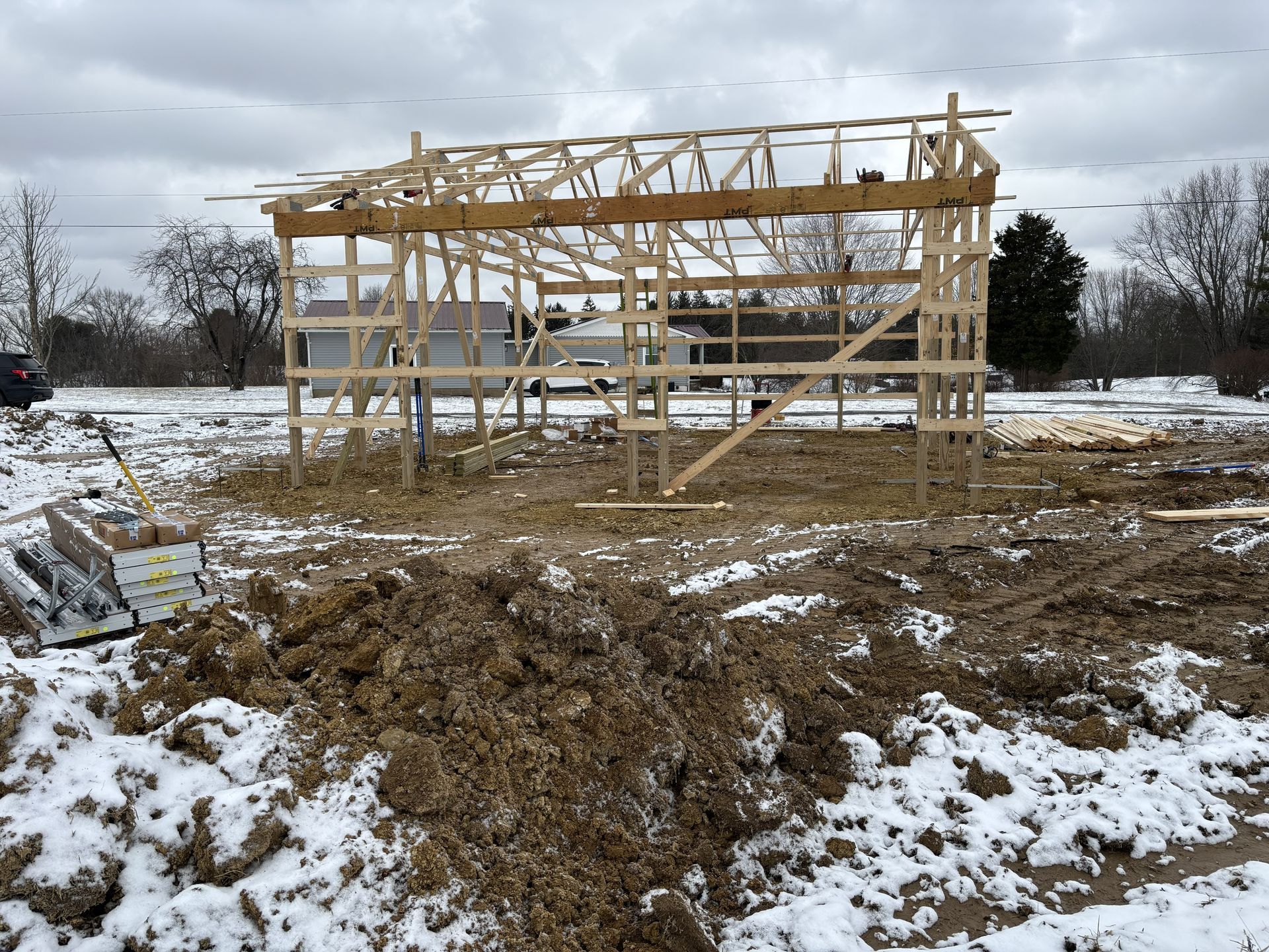Partially built wooden house frame on a snowy, muddy lot under a cloudy sky