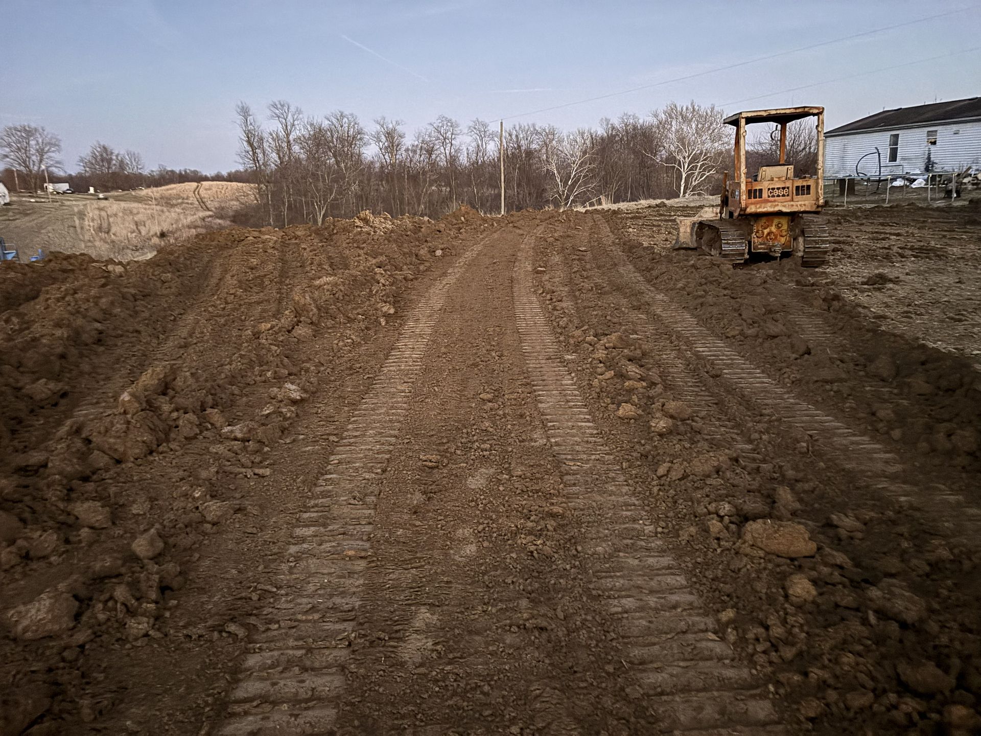 Dirt construction road with a bulldozer on the right and bare trees under a cloudy sky