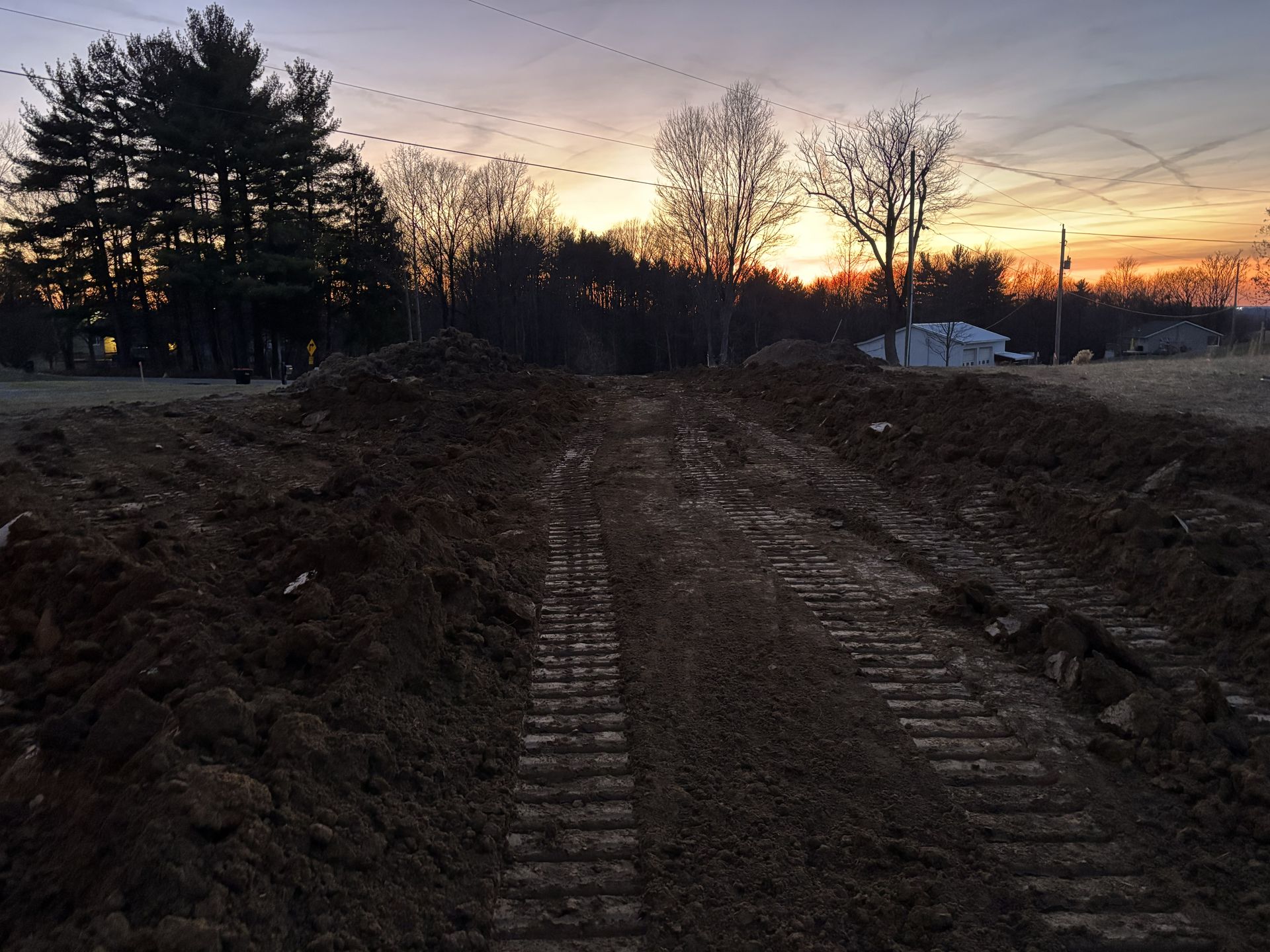 Dirt road under construction at sunset, with trees silhouetted against a glowing sky.