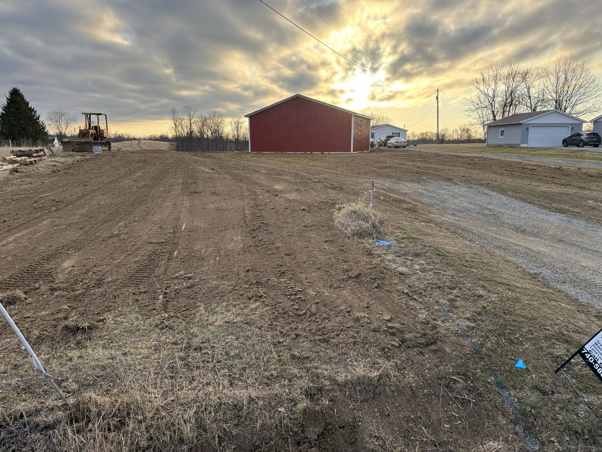 Rural field at sunset with a red barn, dirt track, gravel path, and distant houses and tractor.