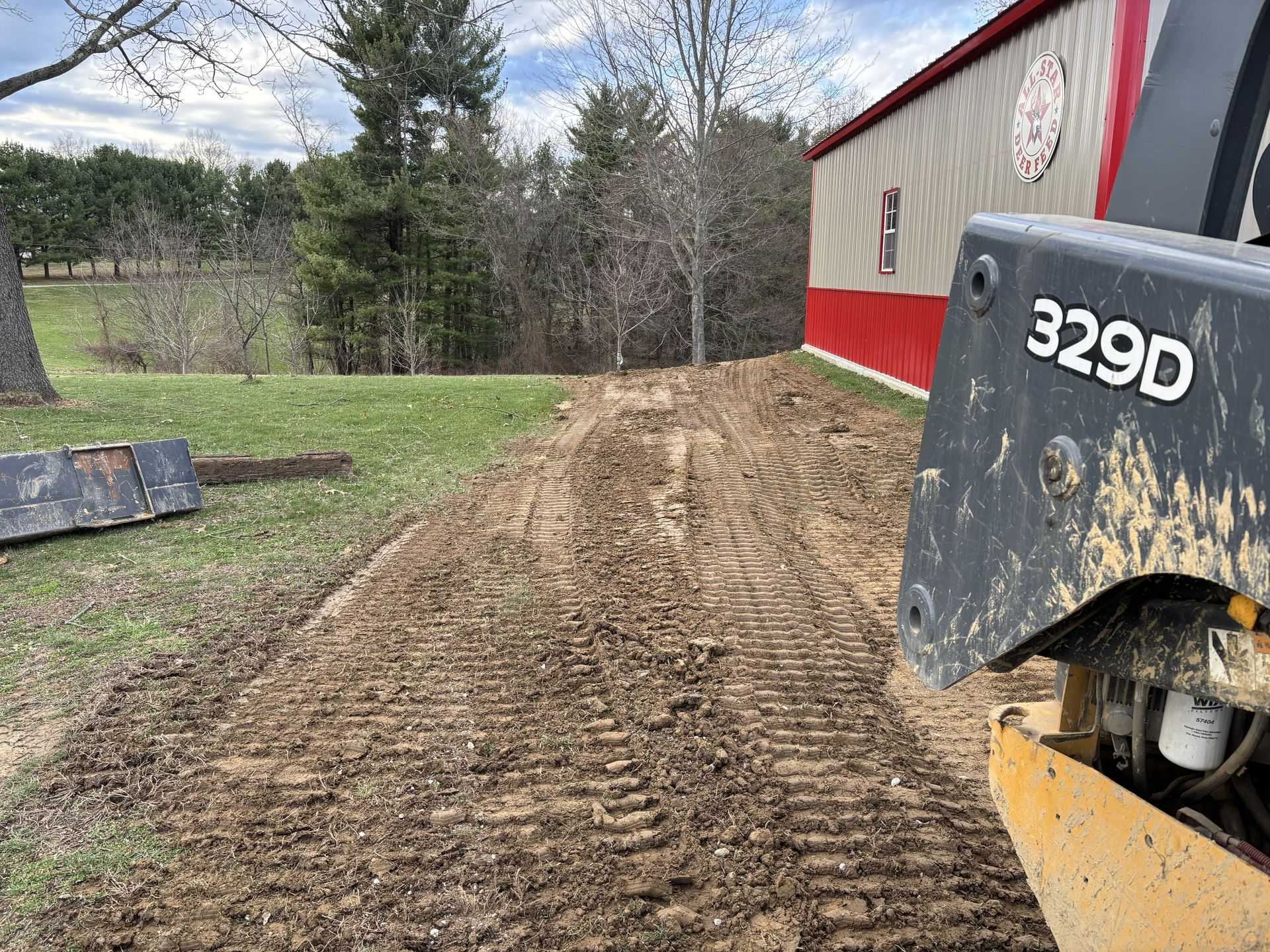 Gravel driveway being graded beside a red barn, with a 329D construction machine in the foreground.