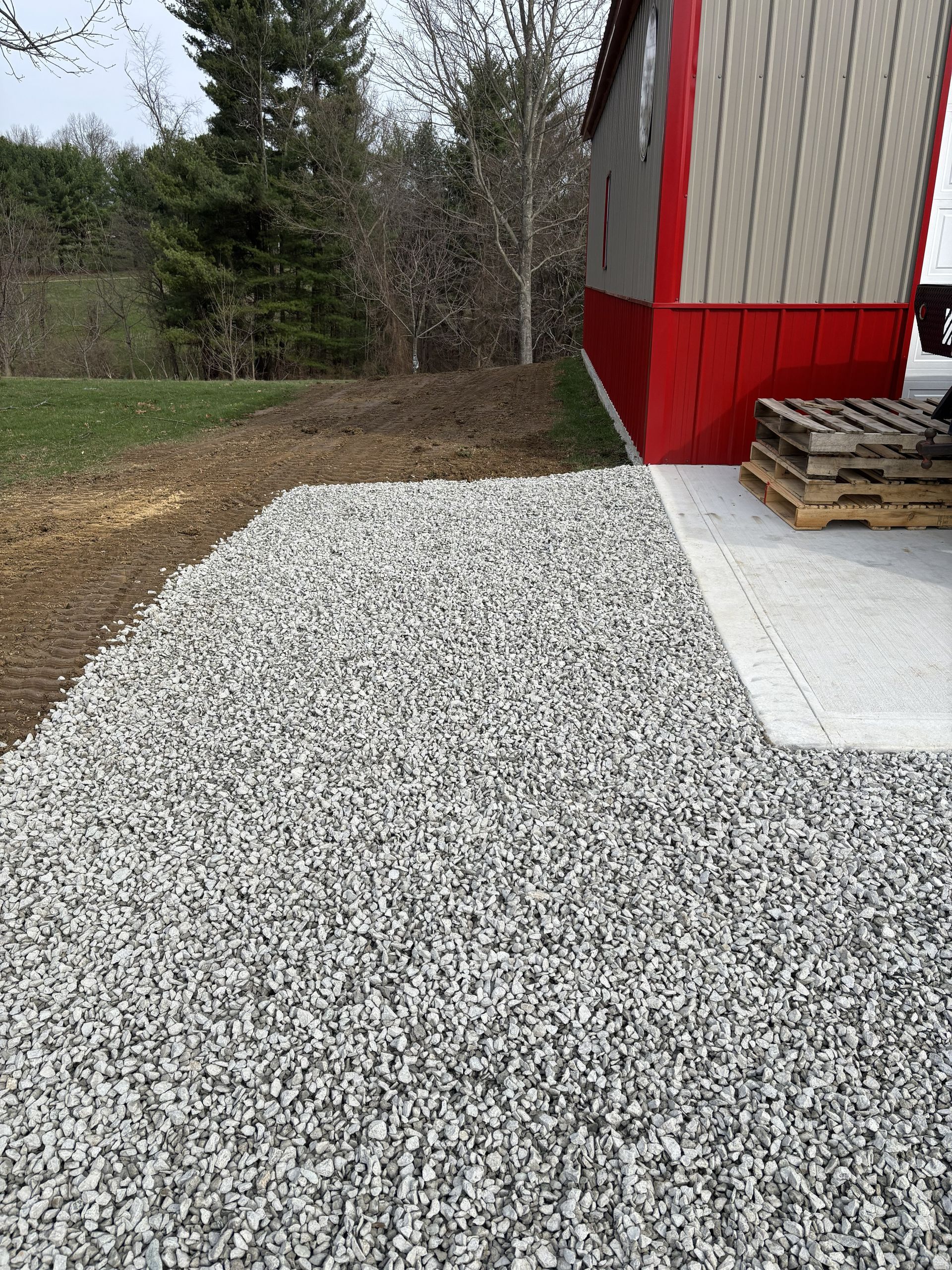 White gravel path beside a red-and-beige building with stacked pallets and trees in the background