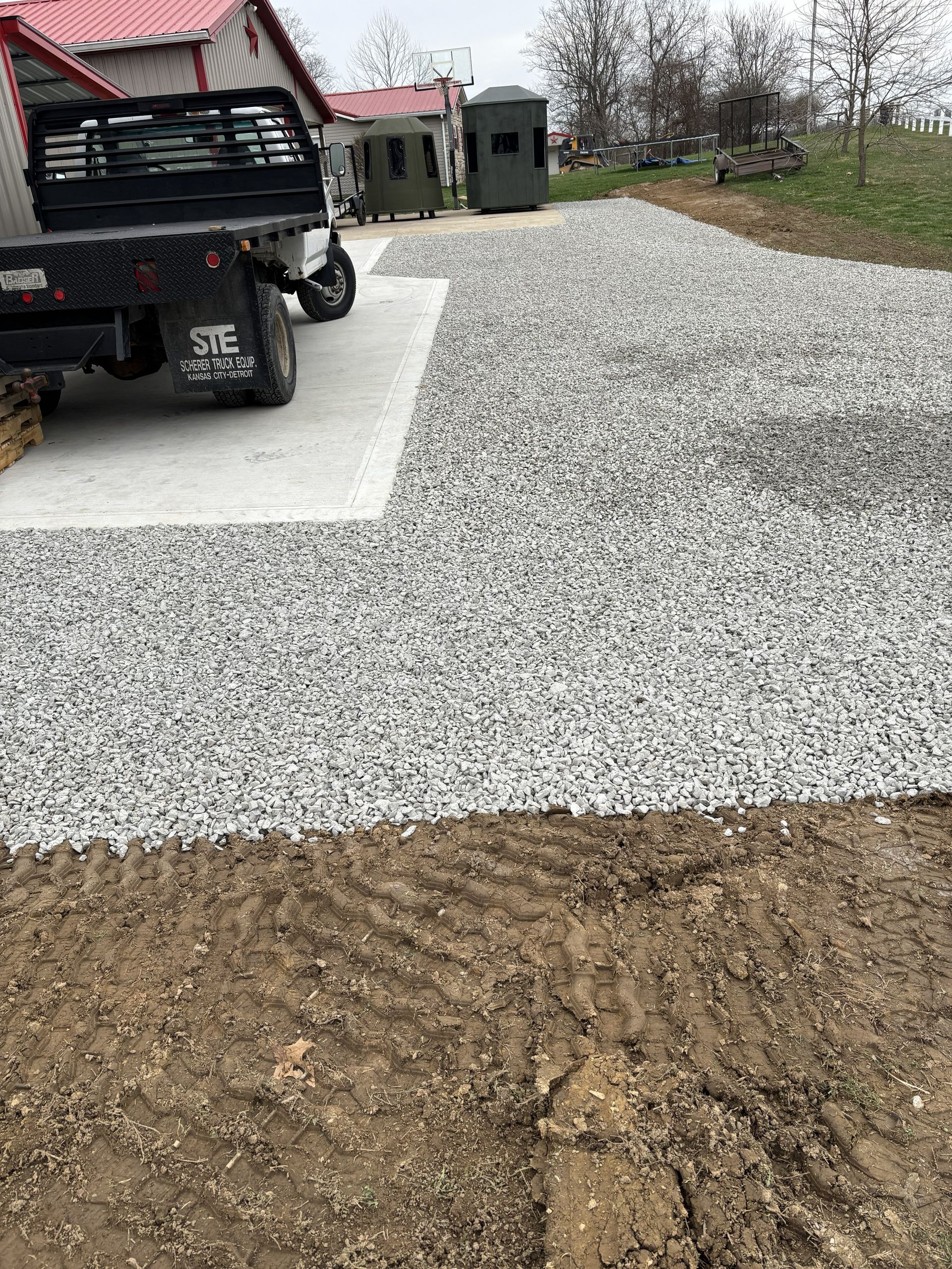 Gravel driveway under construction beside a black utility vehicle, with a dirt foreground and small shed in back