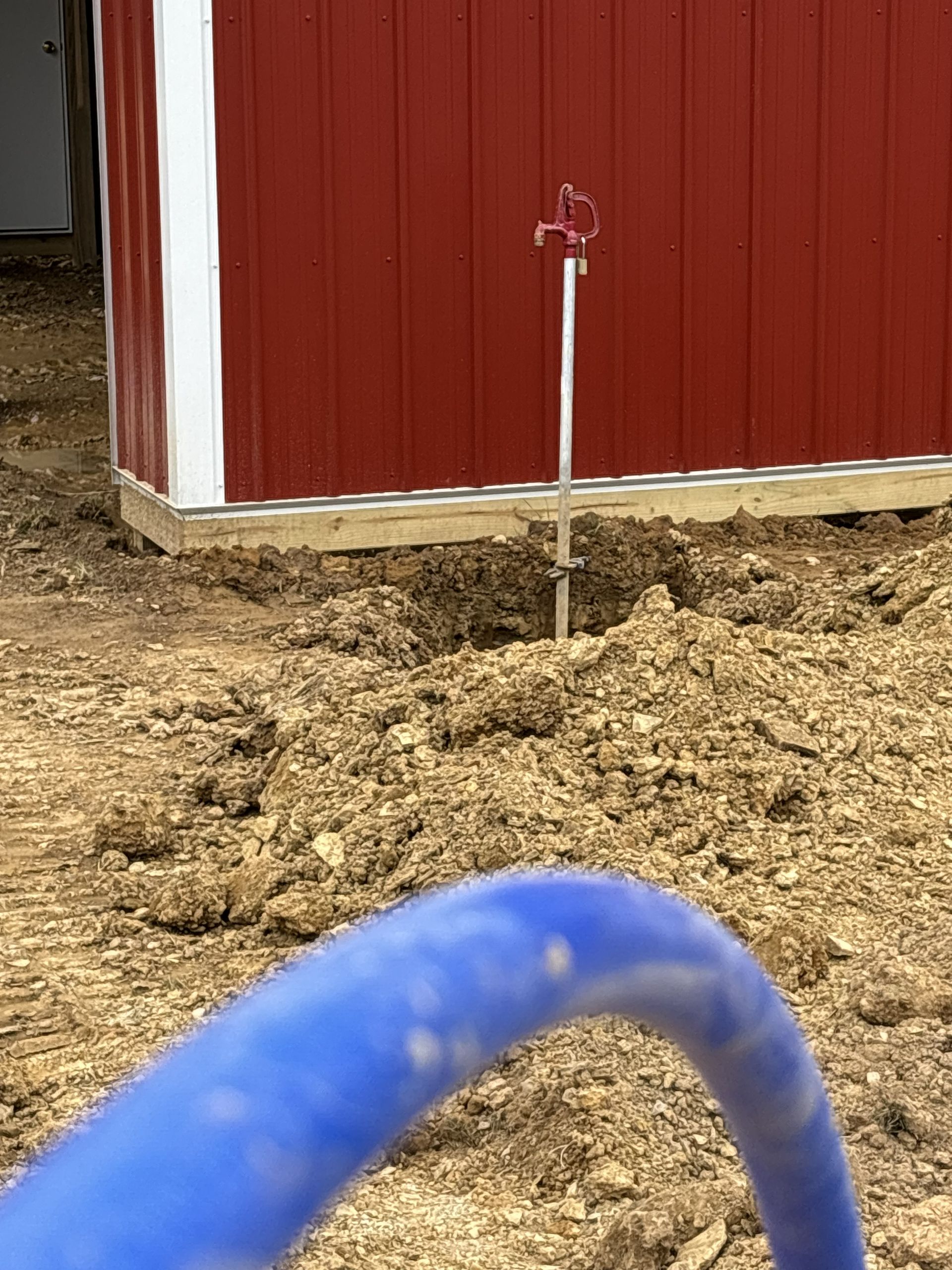 Red barn wall with a small metal stake in muddy ground and a blue hose in the foreground