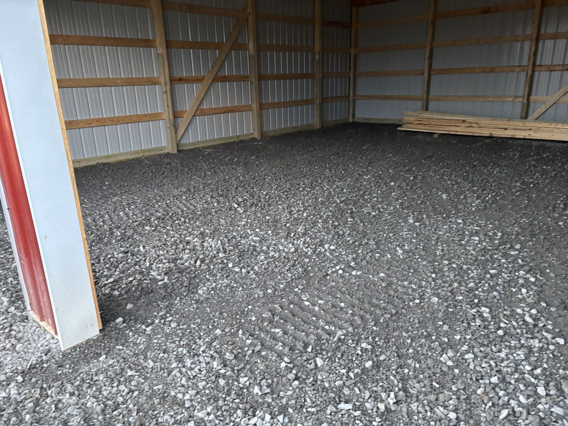 Empty metal barn with gravel floor and stacked lumber along the right wall.