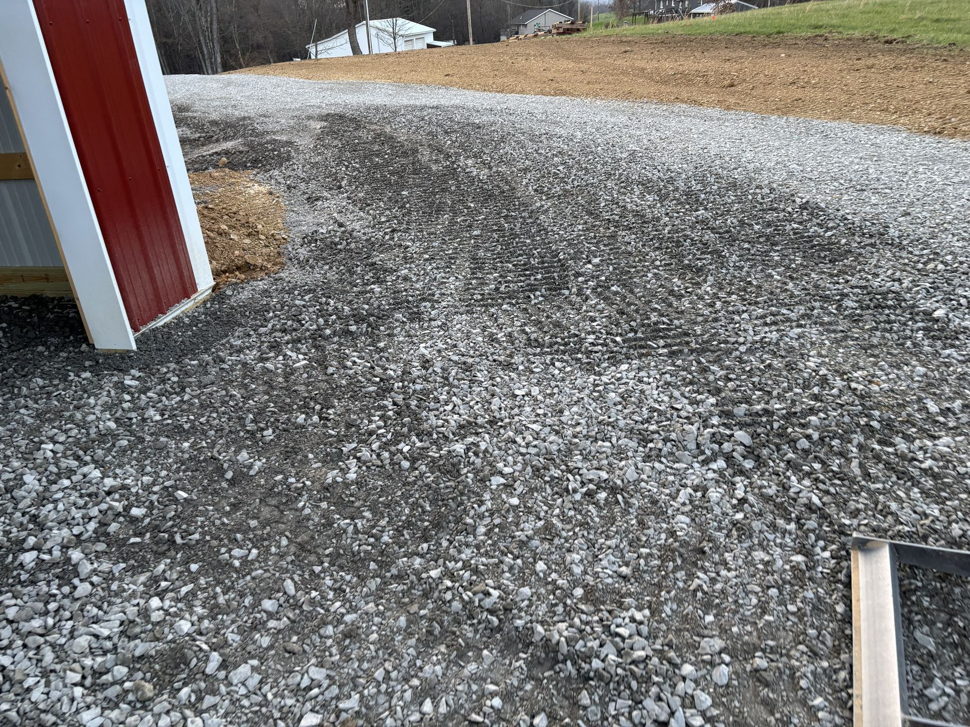 Gravel driveway beside a red building corner, with snow patches and a grassy yard in the background