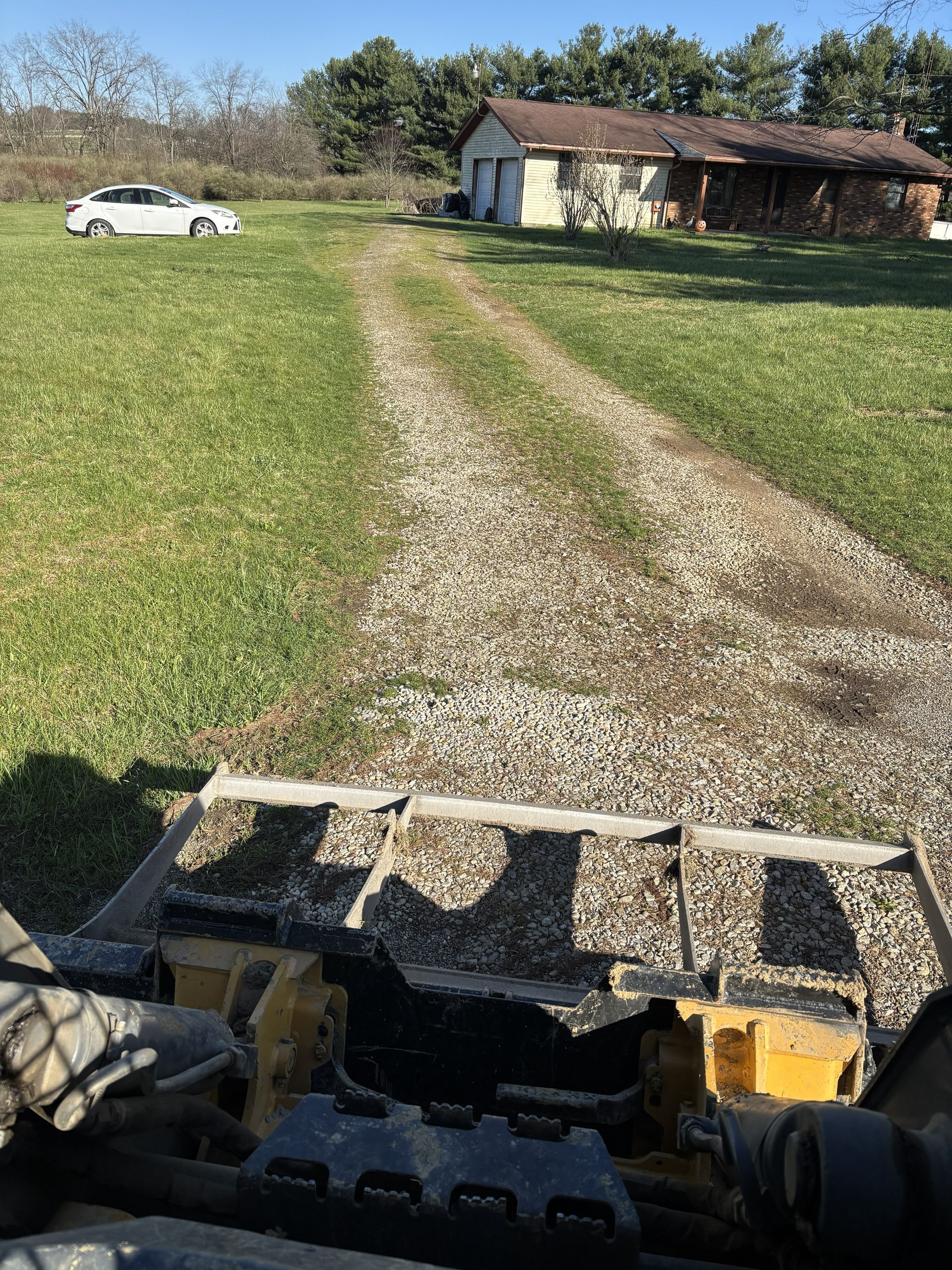 Gravel driveway leading to a small house with a red roof and a white car parked on the lawn.