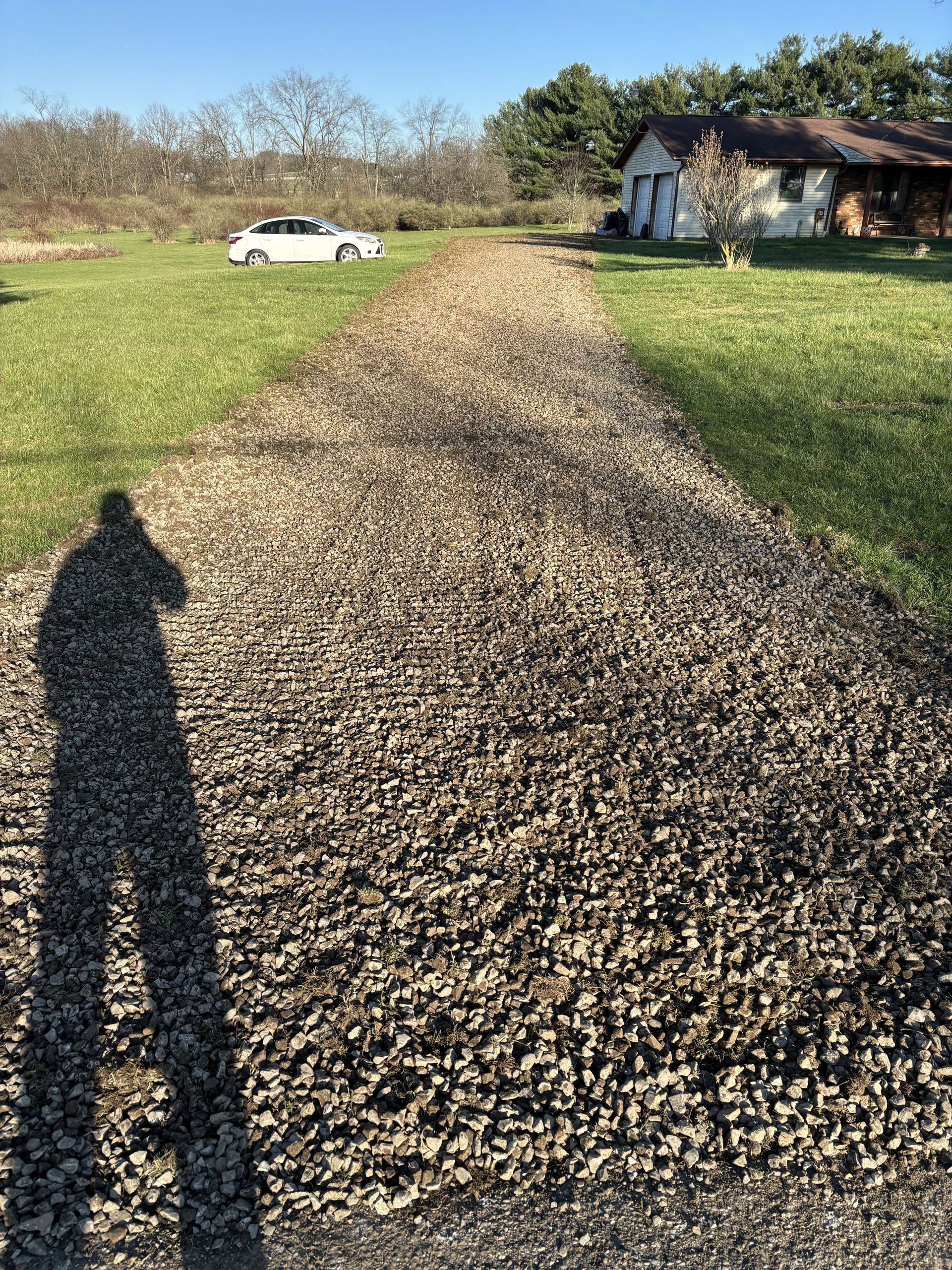 Gravel driveway across a grassy yard toward a white car and house, with a long shadow in the foreground