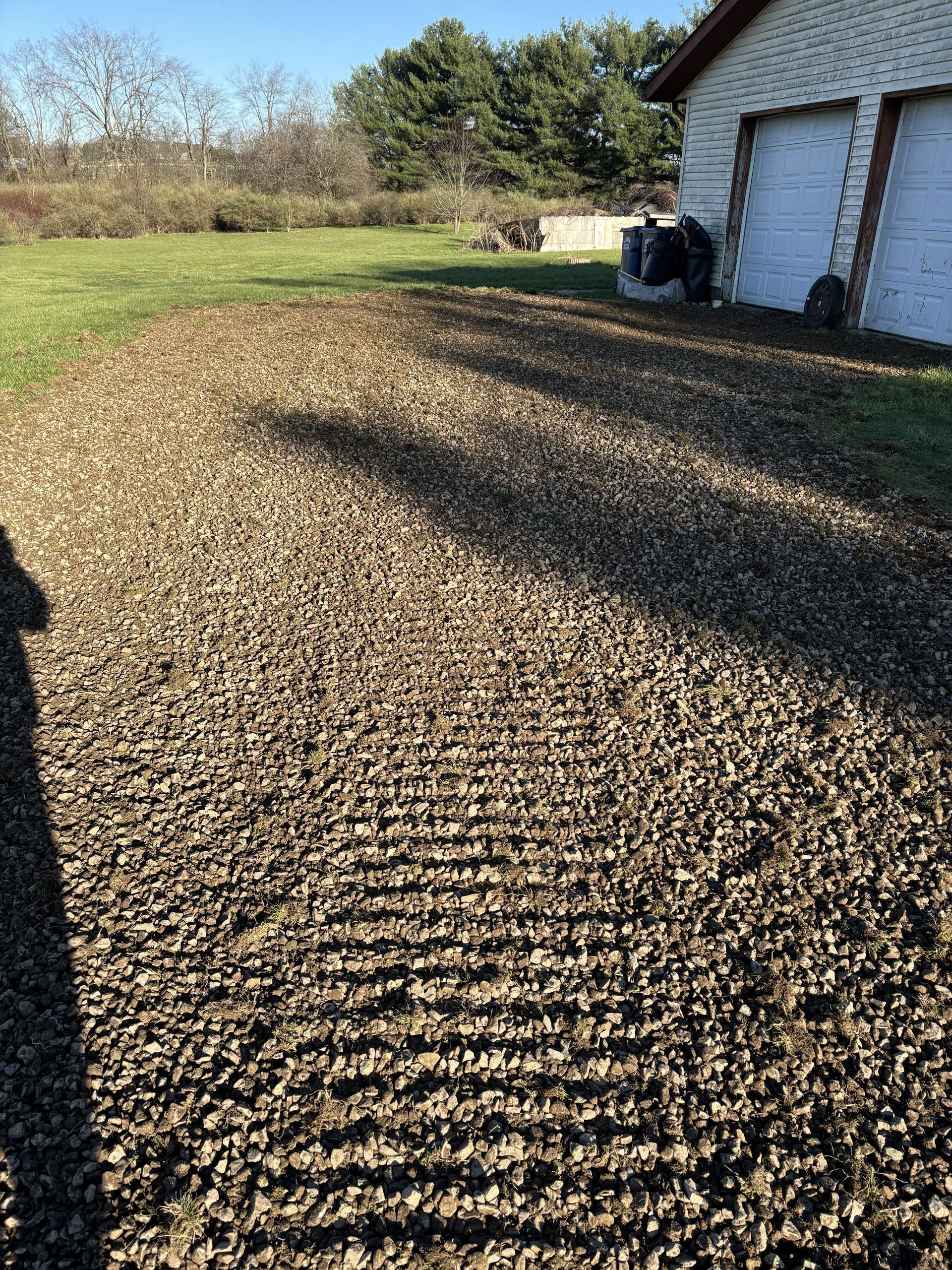 Gravel driveway beside a white garage, with a grassy yard and trees in bright sunlight.