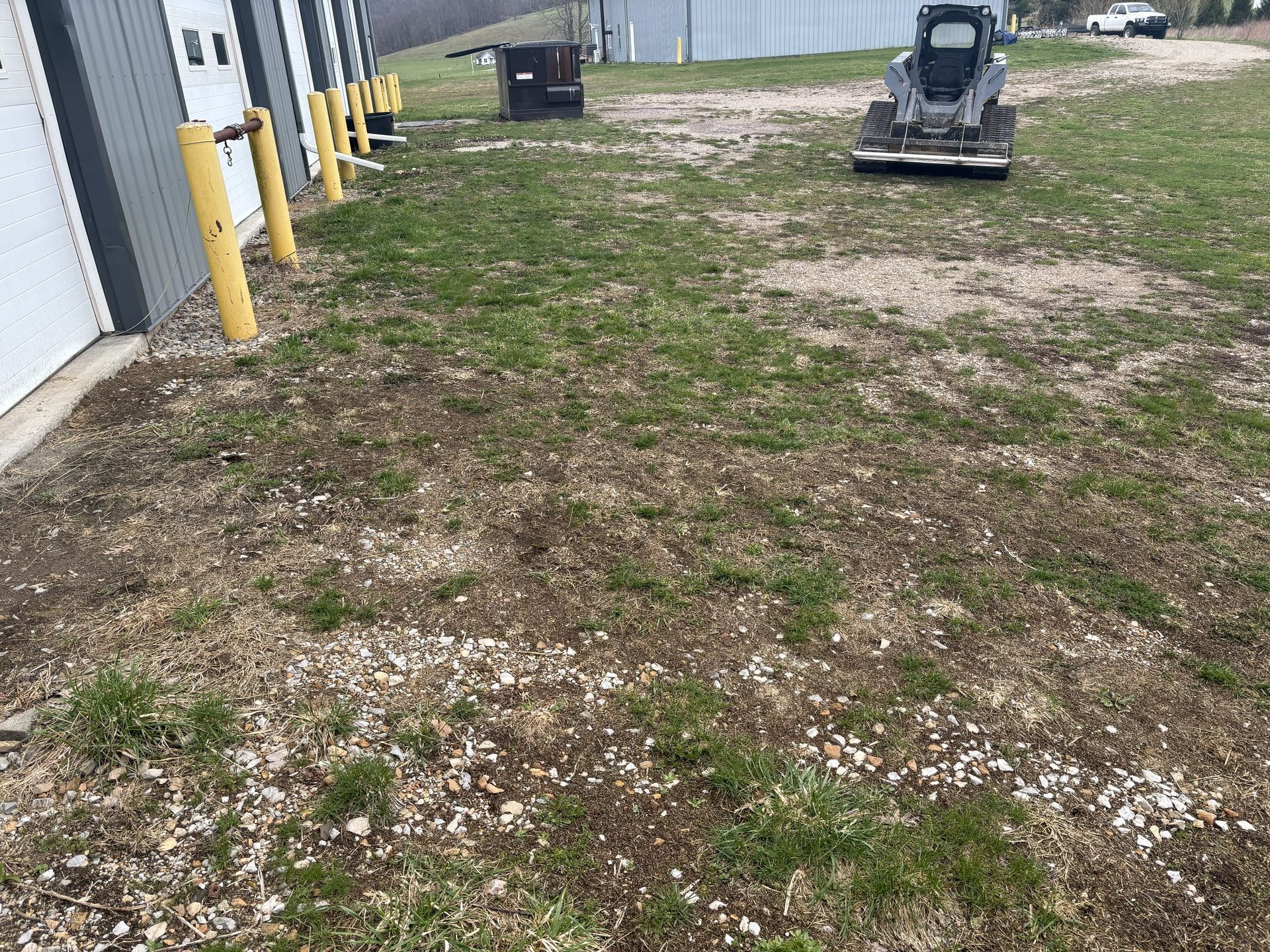 Gravel lot beside a warehouse with yellow bollards and a skid steer in the distance