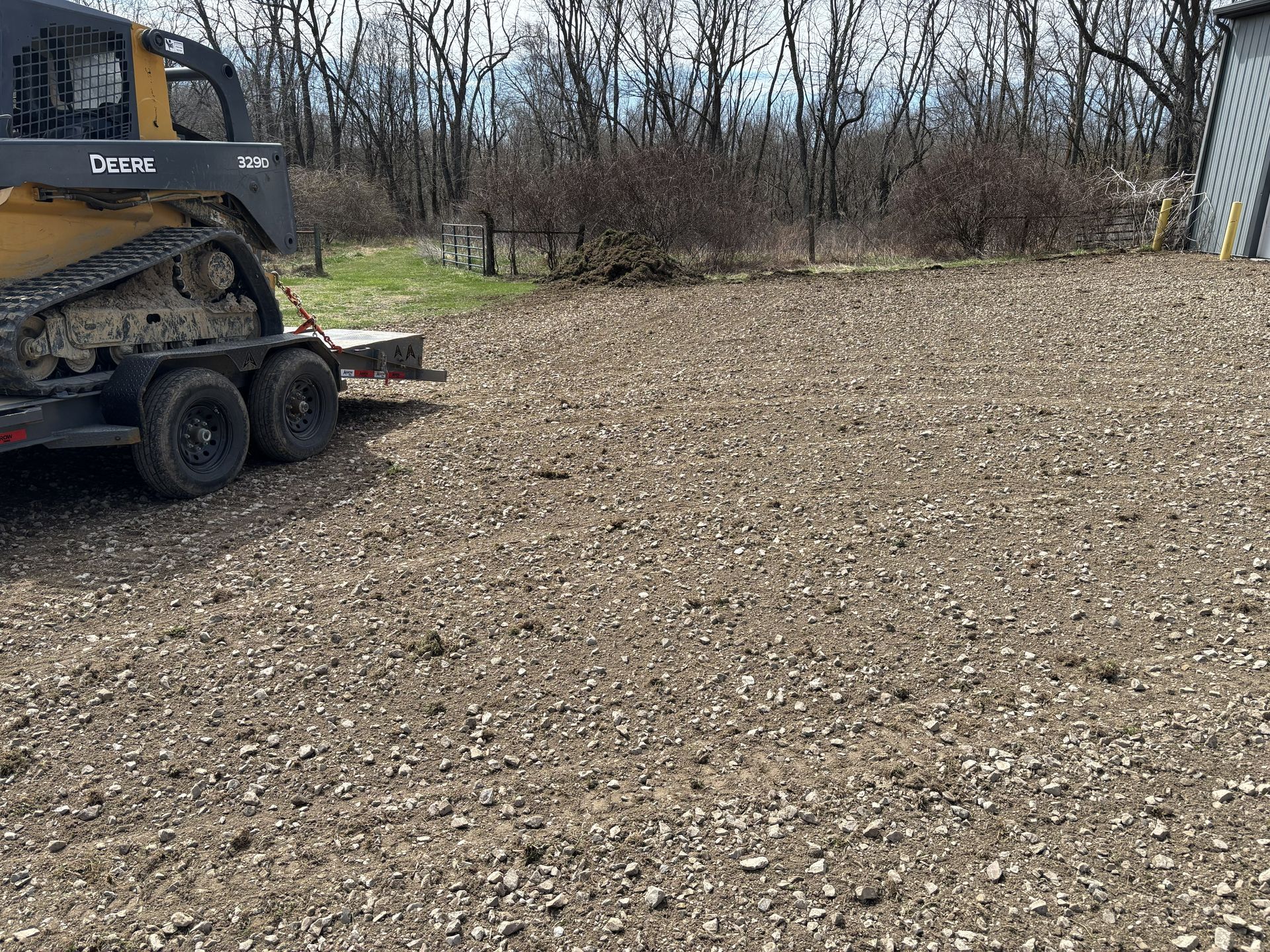 Skid steer loader on gravel lot beside leafless trees and a small building