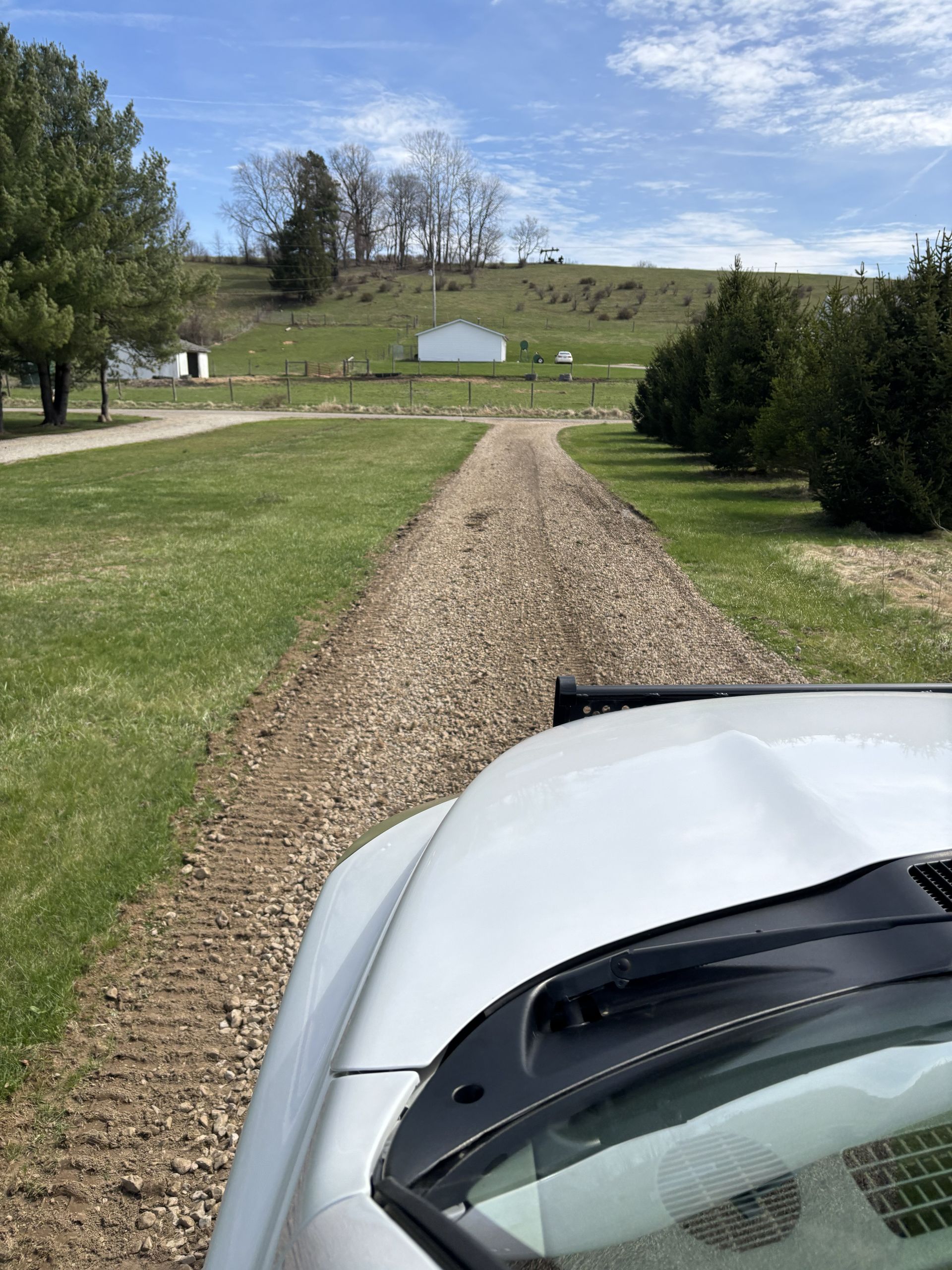 Gravel driveway leading to a white shed in a grassy yard, viewed from a car’s hood under a blue sky.