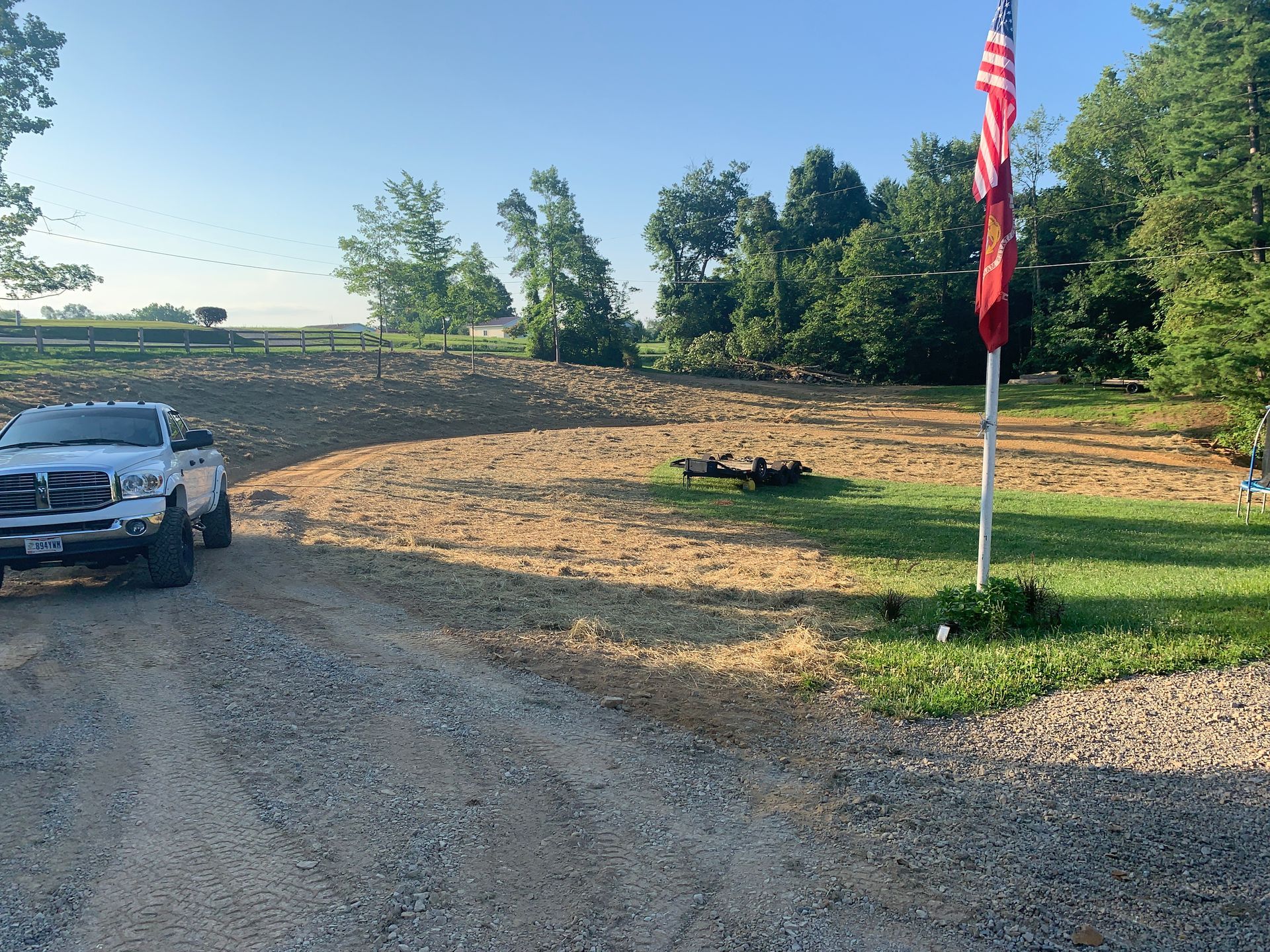 Gravel driveway beside a landscaped lawn, with a parked car at left and an American flag on a pole at right.