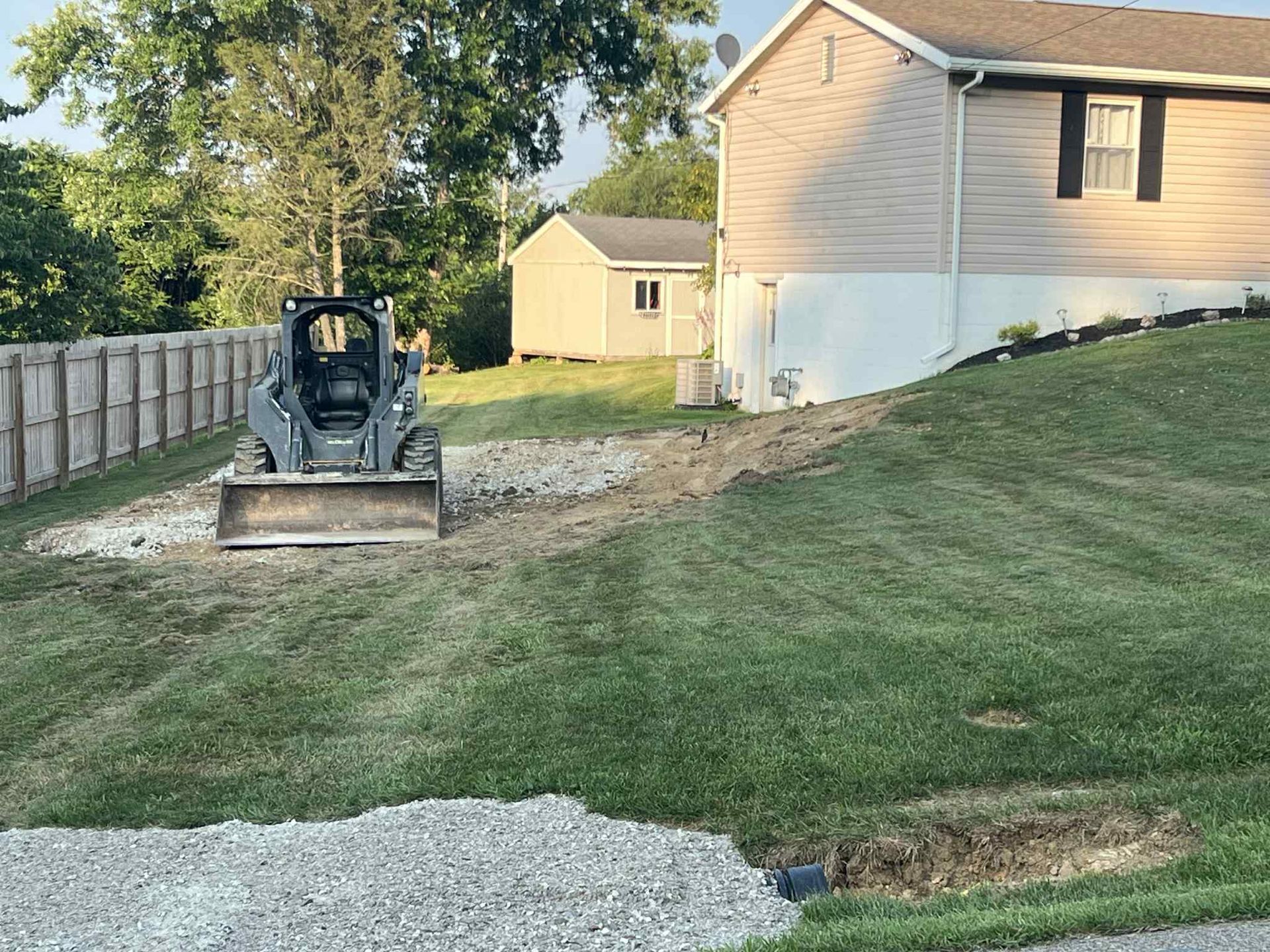 Mini excavator leveling a dirt patch beside a suburban house and fence in a backyard.