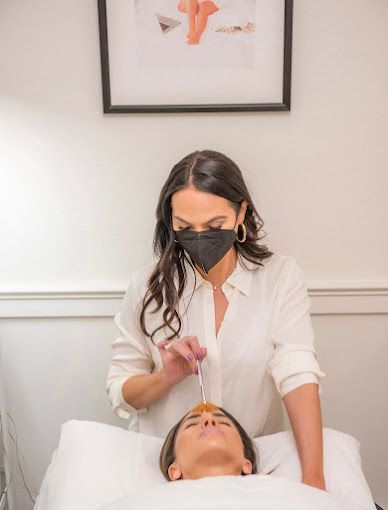 A woman wearing a mask is applying a facial treatment to a woman laying on a bed.