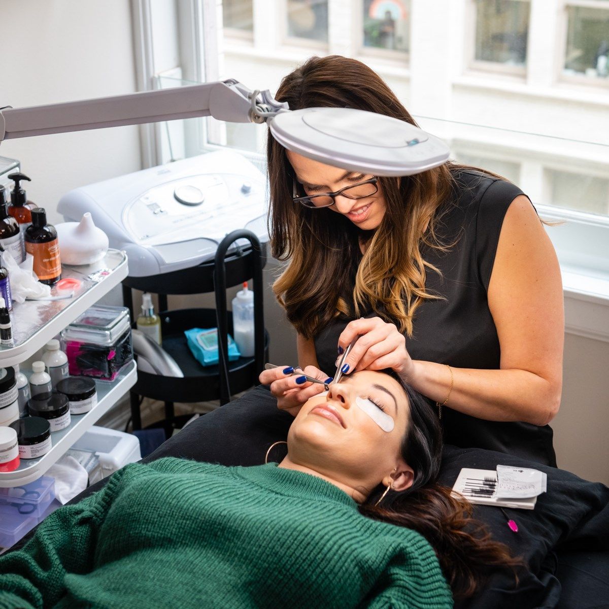 A woman in a green sweater is getting her eyelashes done by a woman in a black shirt.
