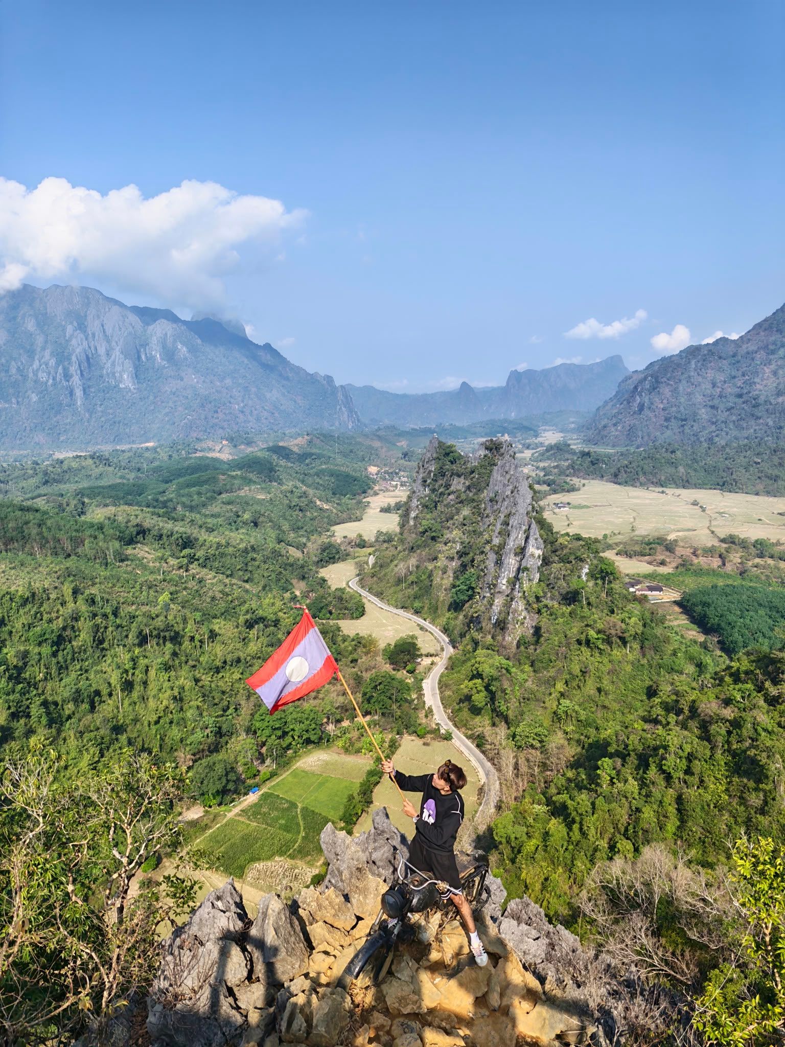 Hiker on rocky mountain ridge above lush valley, with a red-white flag and distant peaks under blue sky
