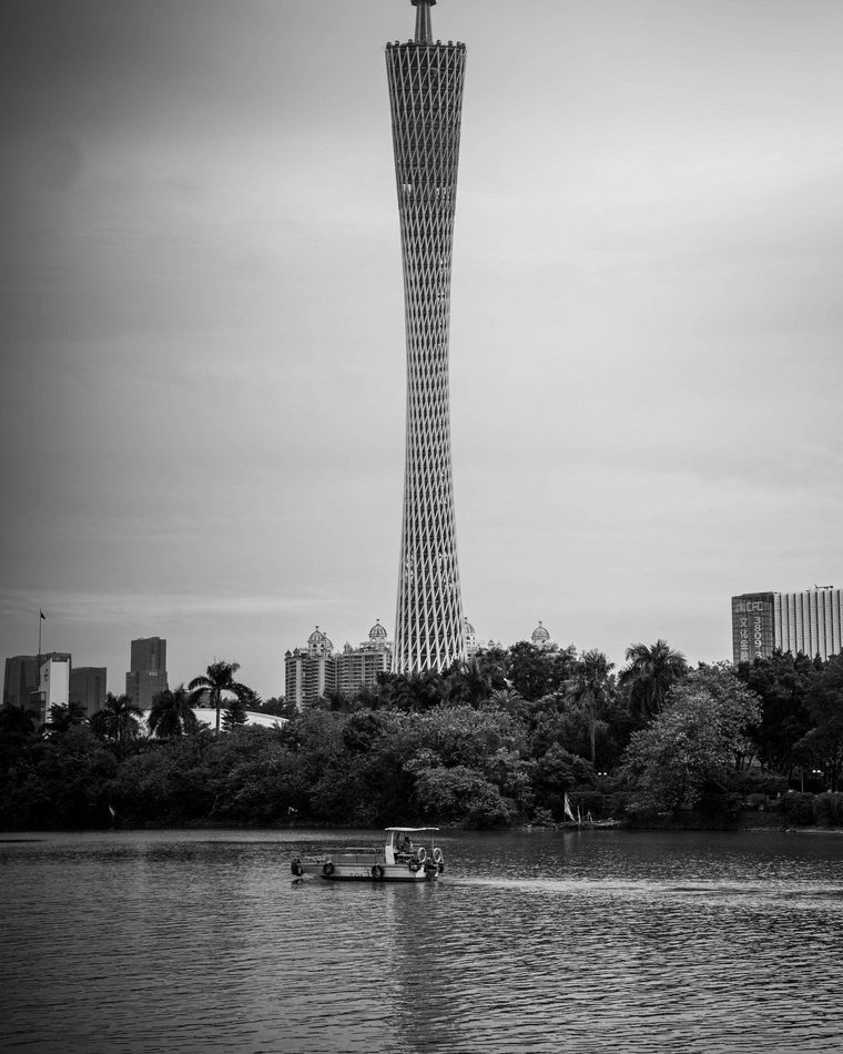 Black-and-white waterfront skyline with a tall twisting tower rising behind trees and boats on the water