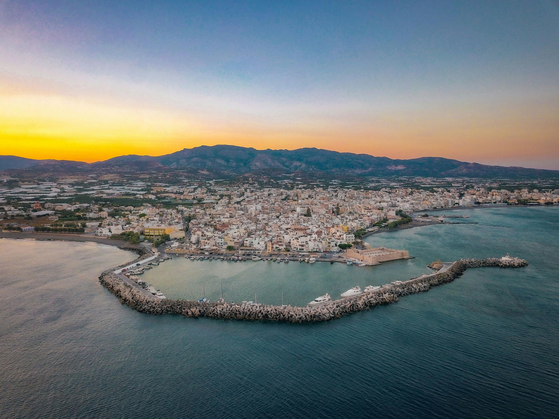 Aerial view of a coastal city at sunset with a curving harbor, rocky breakwater, and mountains in the background