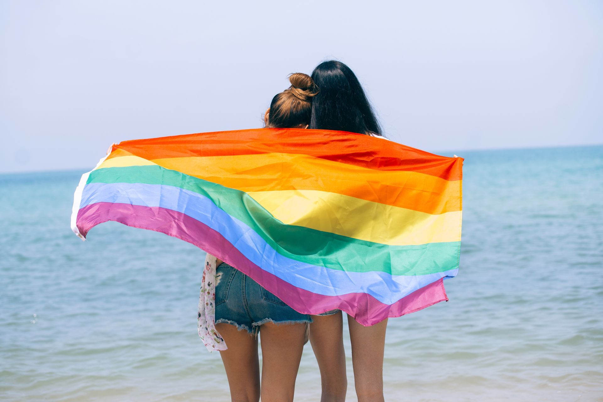 Two people on a beach holding a rainbow Pride flag out to the sea