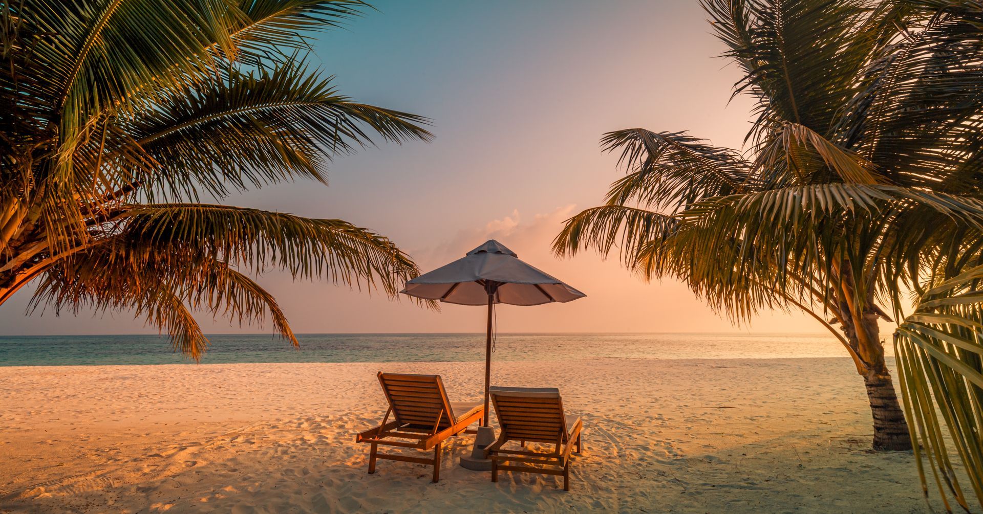 Two beach chairs under an umbrella at sunset, framed by palm trees on a sandy beach