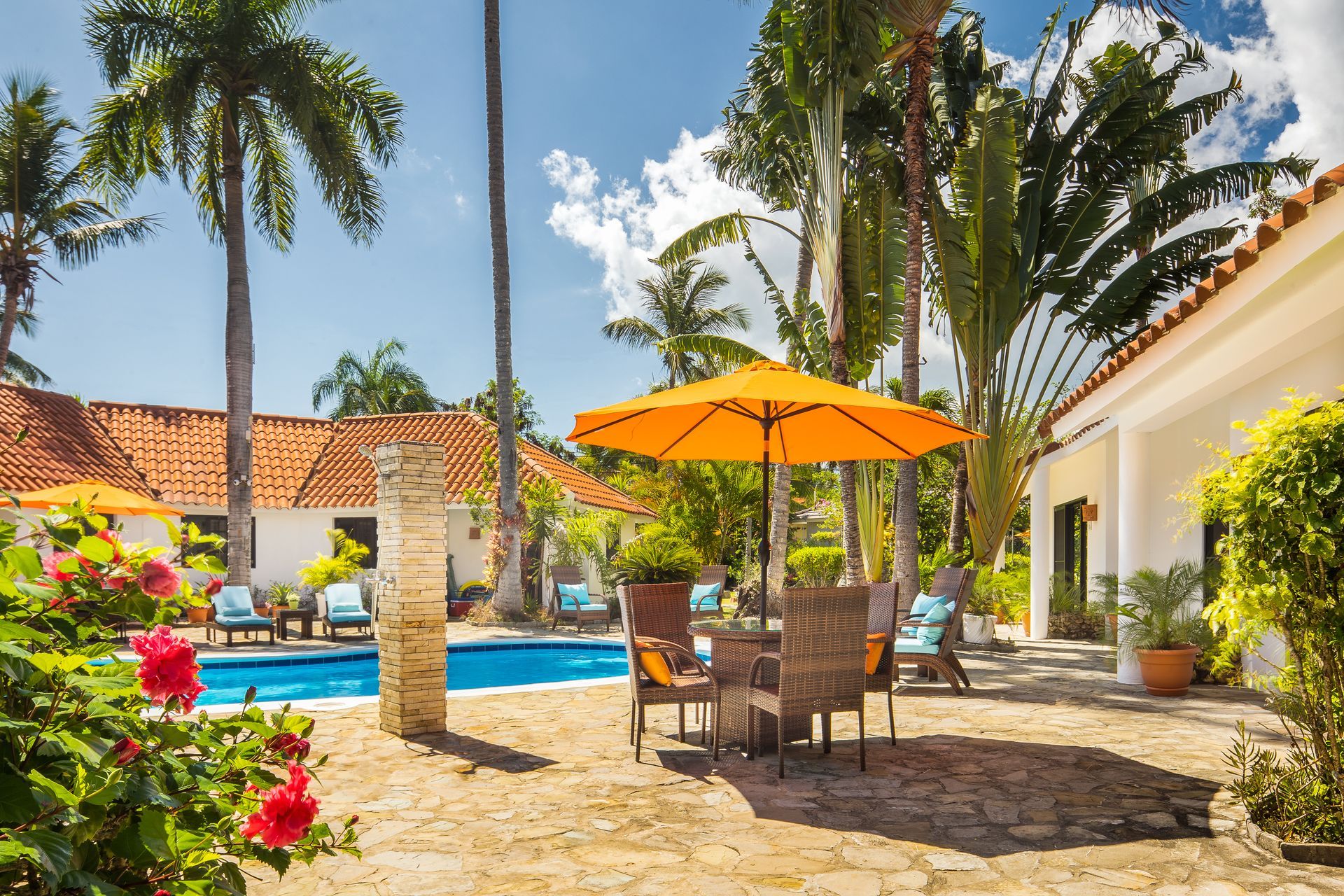 Tropical courtyard with pool, dining set under orange umbrella, palm trees, and white buildings.