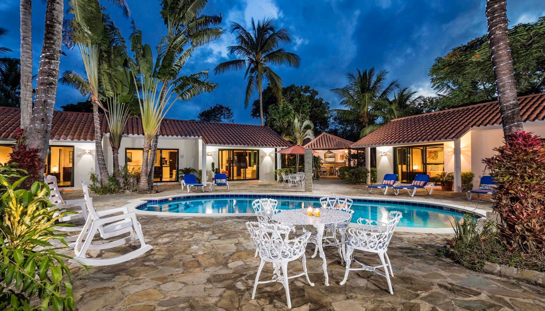 Poolside villas with white furniture, blue pool, palm trees, and dusk lighting.