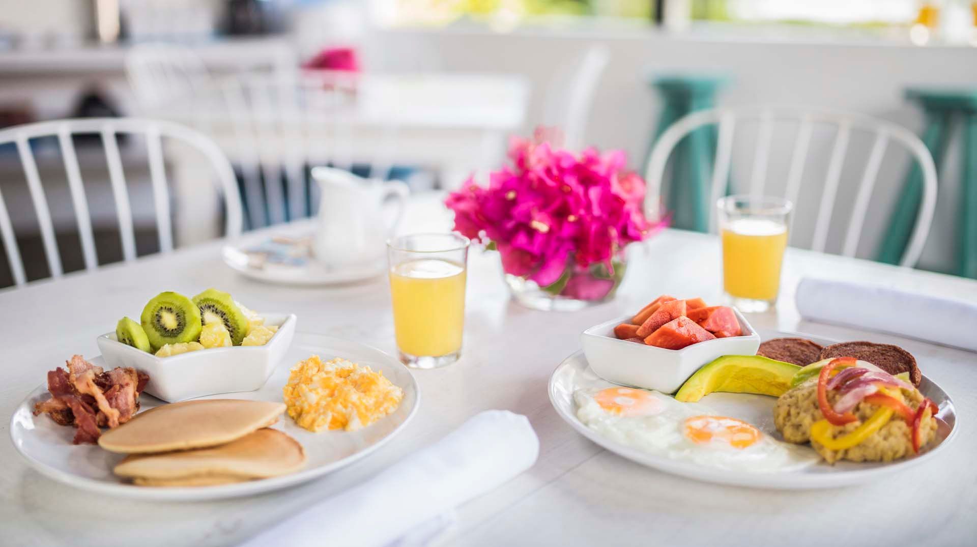 Breakfast spread on a white table: pancakes, eggs, fruit, juice, and flowers. Bright, sunny setting.
