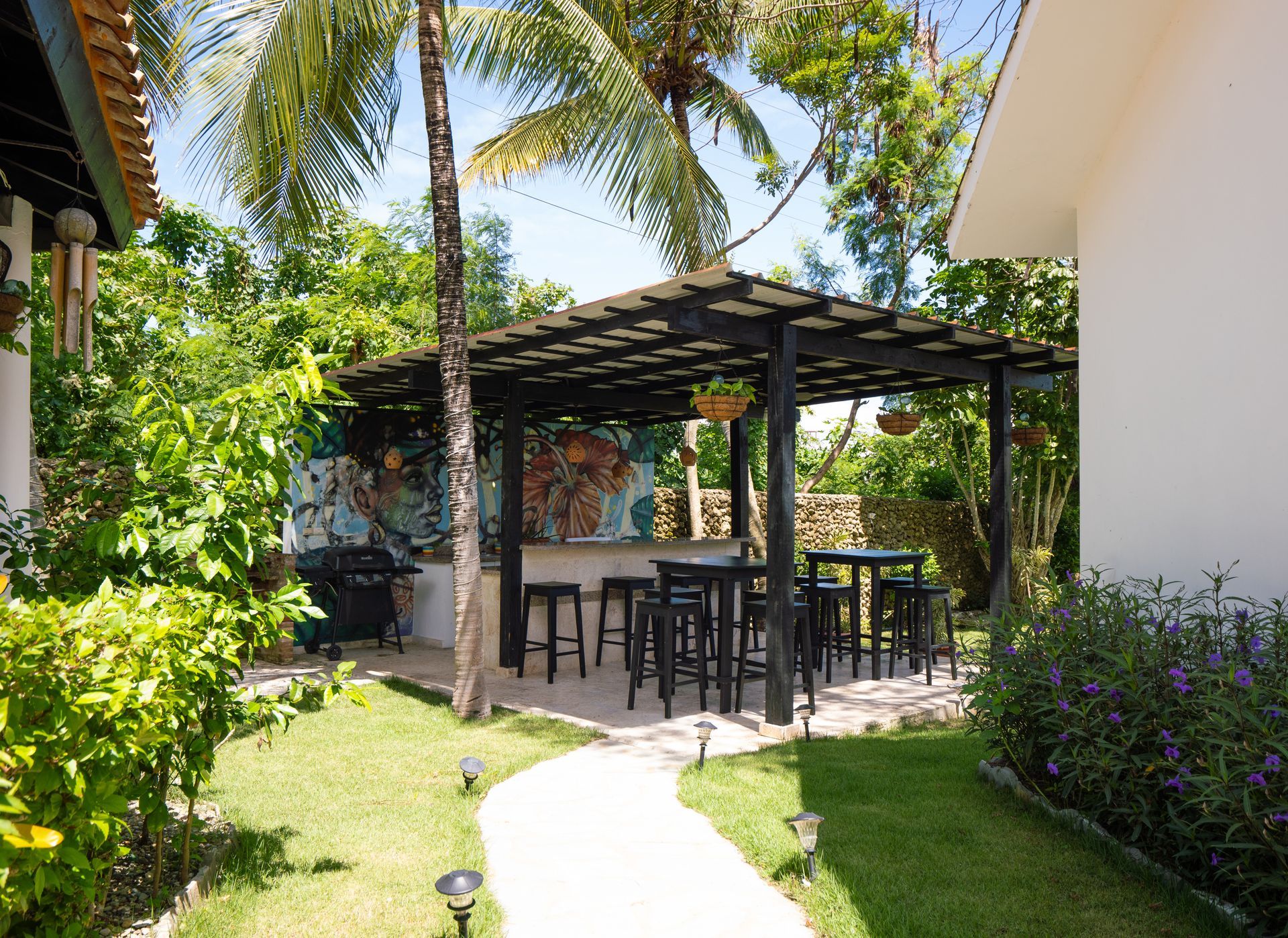 Outdoor bar with black stools and table under a pergola in a lush green yard.