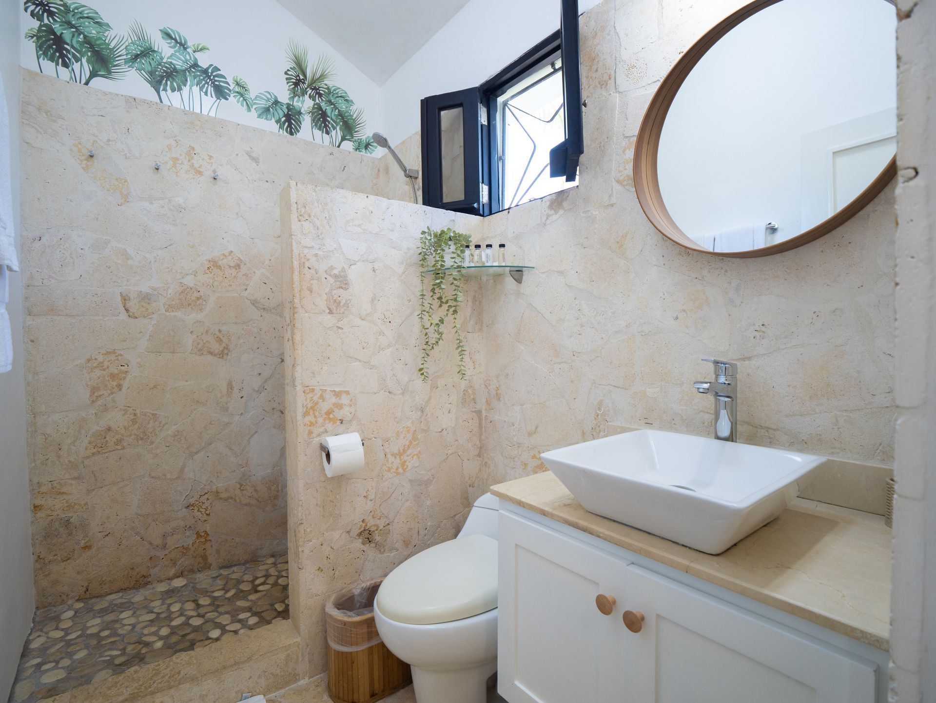Bathroom with stone shower, vanity, toilet, and round mirror. Window above.
