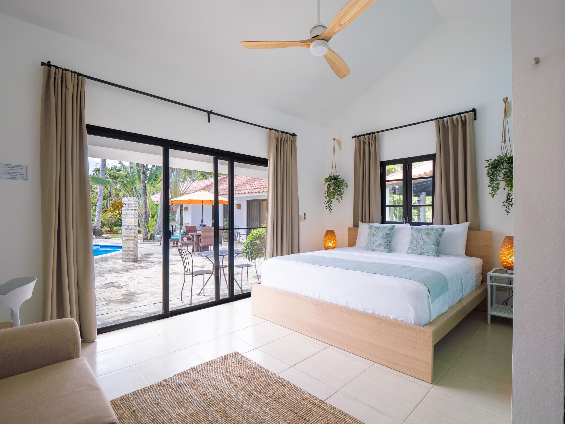 Bedroom with open sliding doors to a pool and patio. Light wood bed frame, neutral curtains, and hanging plants.
