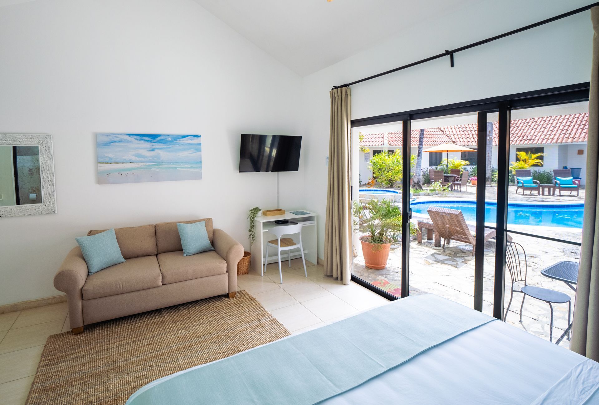 Bedroom with pool view: beige couch, blue accents, TV, desk, and sliding glass door.