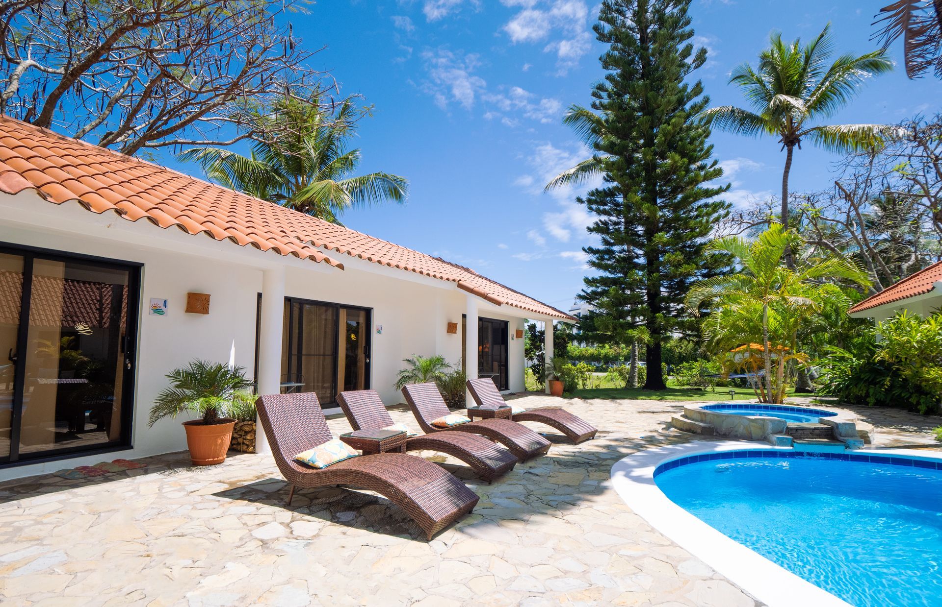 A sunny villa with a pool and lounge chairs. White walls, orange tile roof, palm trees.