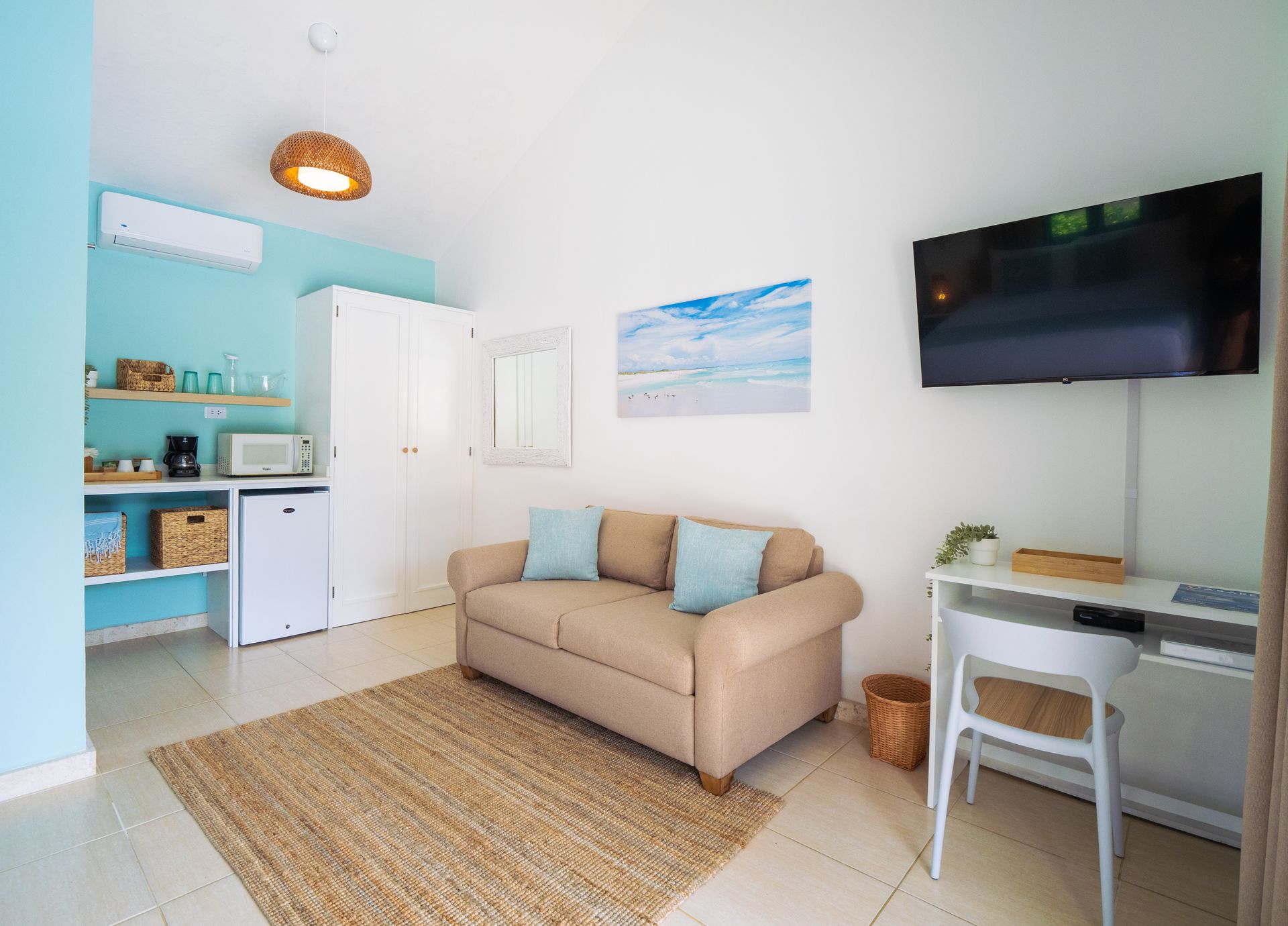 Bright, airy living space with a beige sofa, desk, kitchenette, and wall-mounted TV. Blue and white decor.