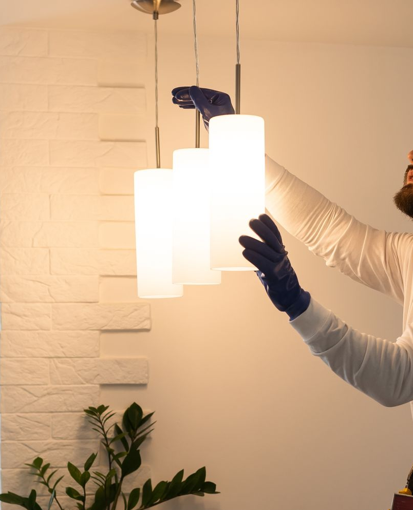 A person wearing blue gloves adjusts three cylindrical, glowing pendant lights in a room with a white brick wall.