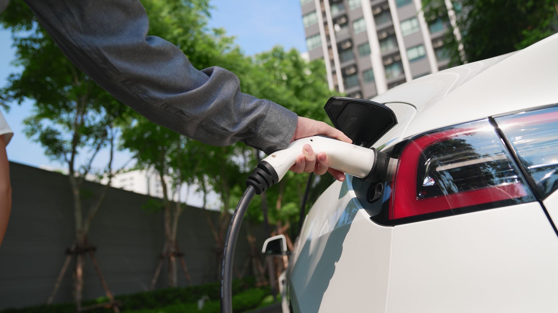 A person charging a white electric vehicle at an outdoor station near trees and a building.