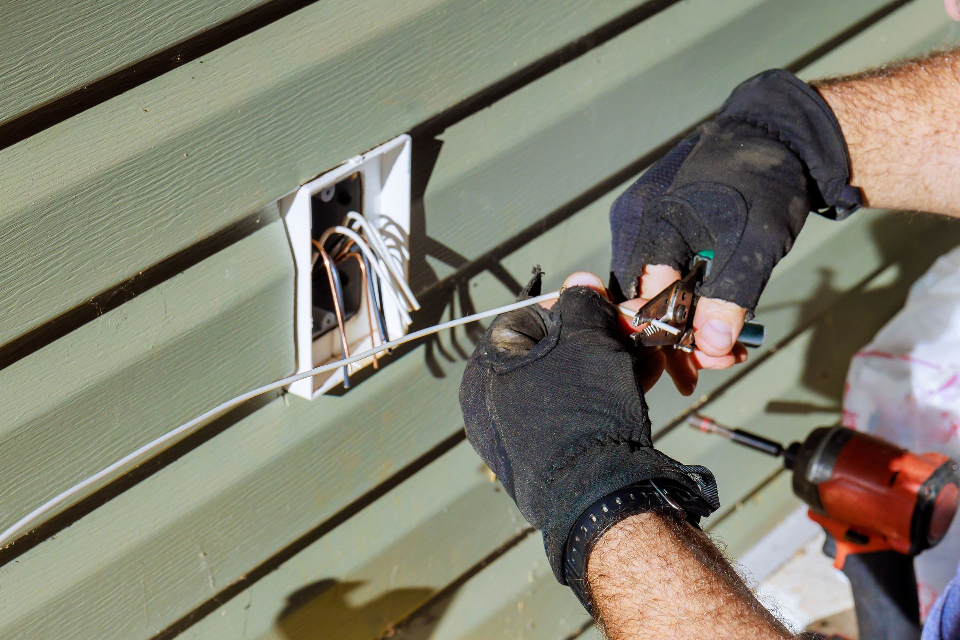 A person wearing work gloves cuts an electrical wire near an open exterior wall junction box on green siding.