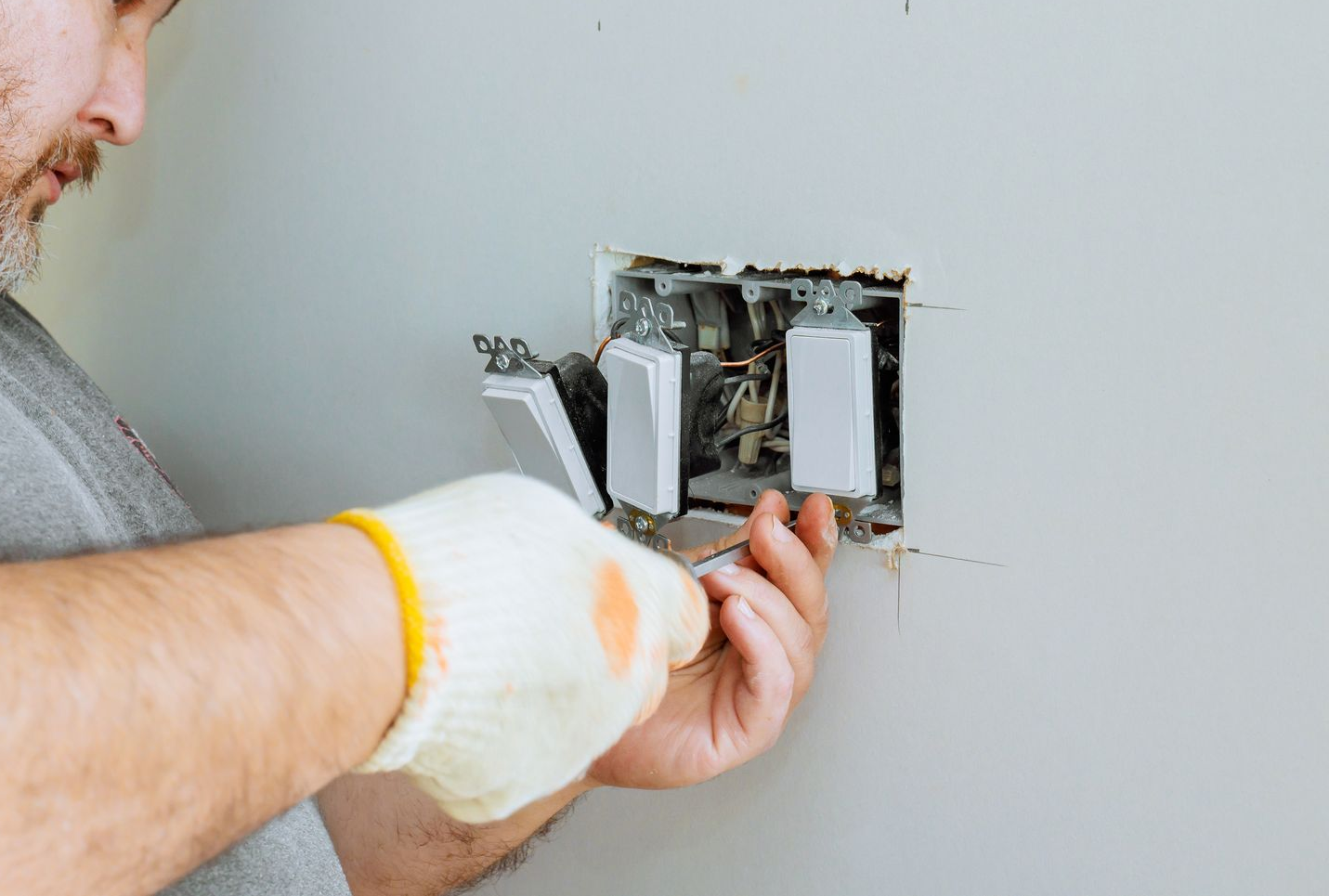 A person wearing gloves works on an electrical wall switch assembly with multiple toggles.