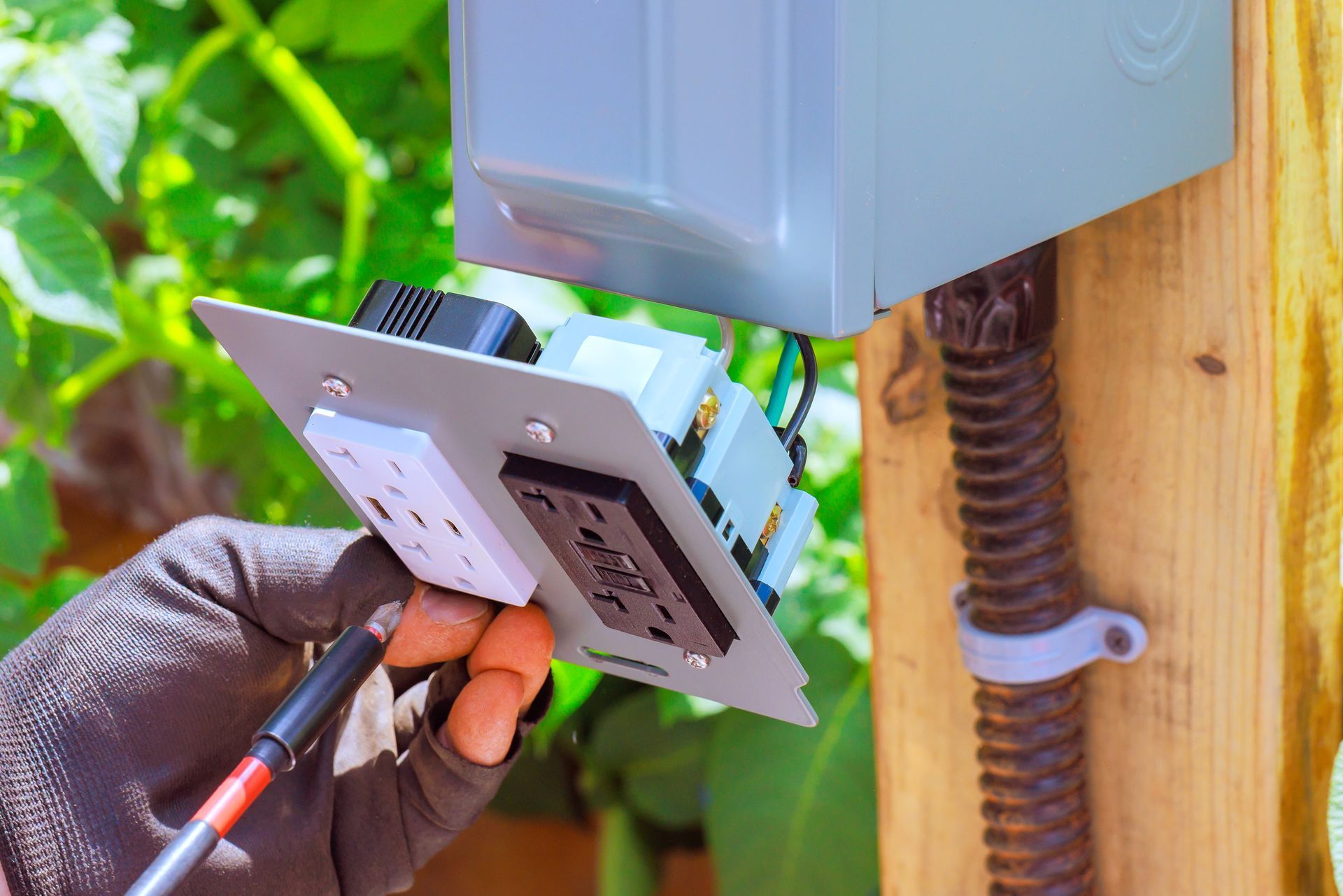 A gloved hand uses a tool to install electrical outlets into an outdoor junction box mounted on a wooden post.