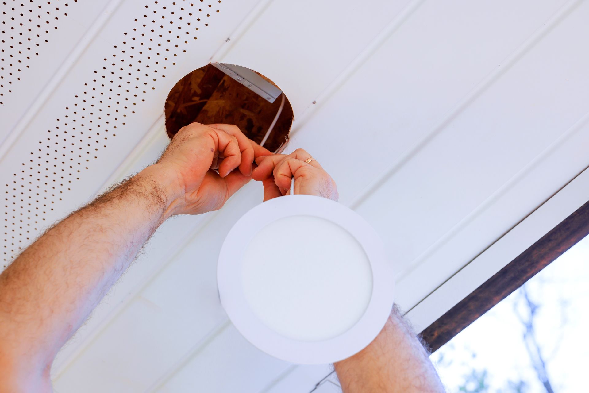 A person installing a circular recessed light fixture into a white soffit ceiling.