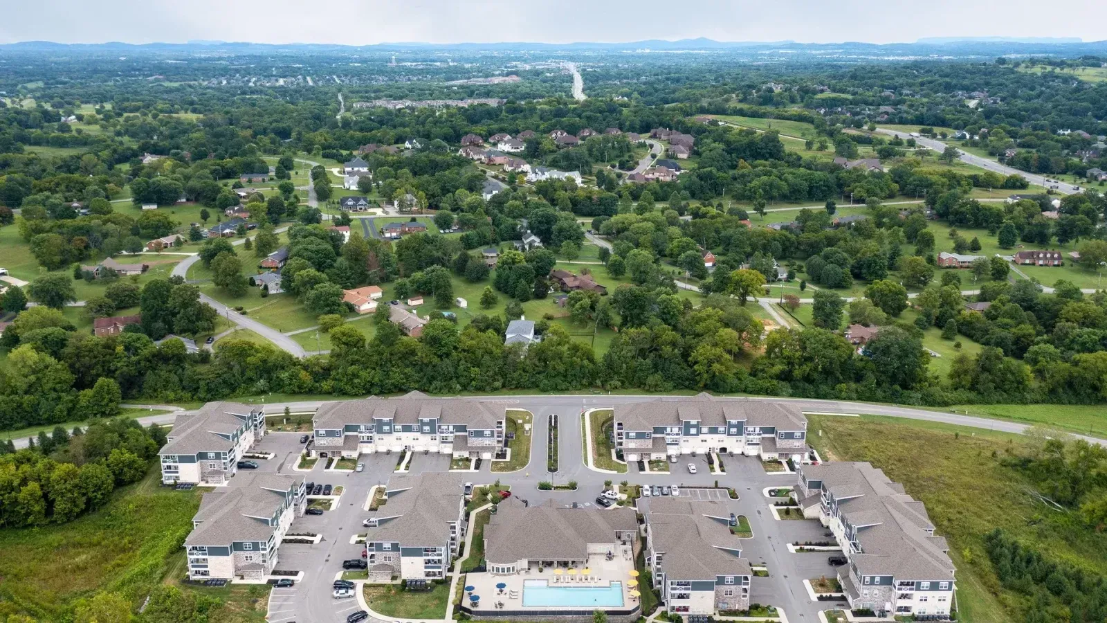 Aerial view of a suburban apartment complex with parking lots, trees, and a green residential neighborhood beyond