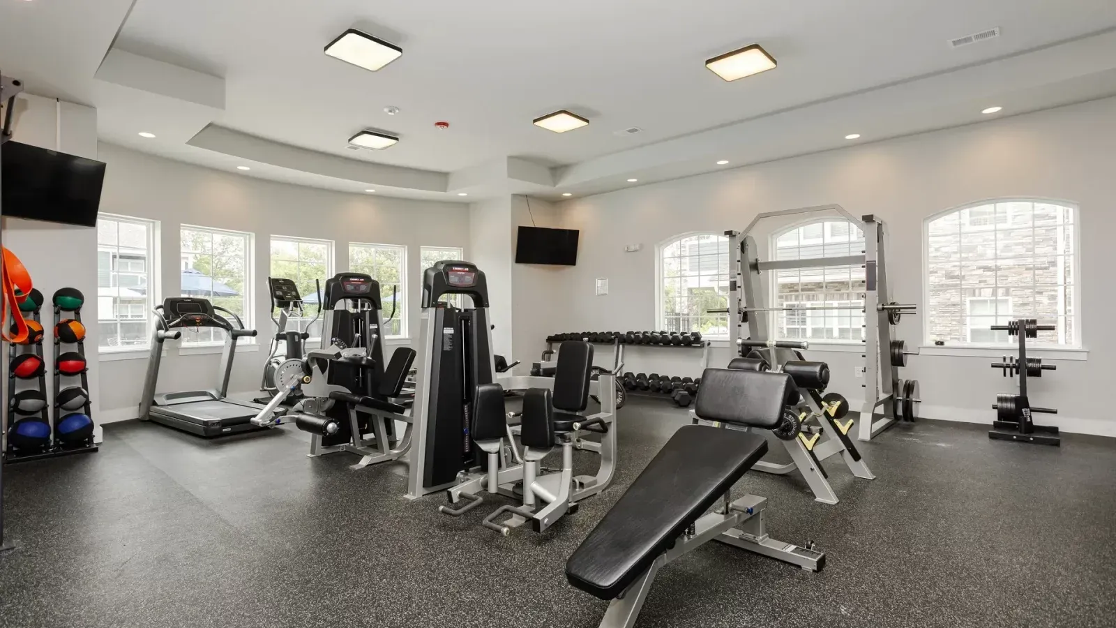 Bright gym with weight machines, benches, free weights, and large windows in a spacious white room