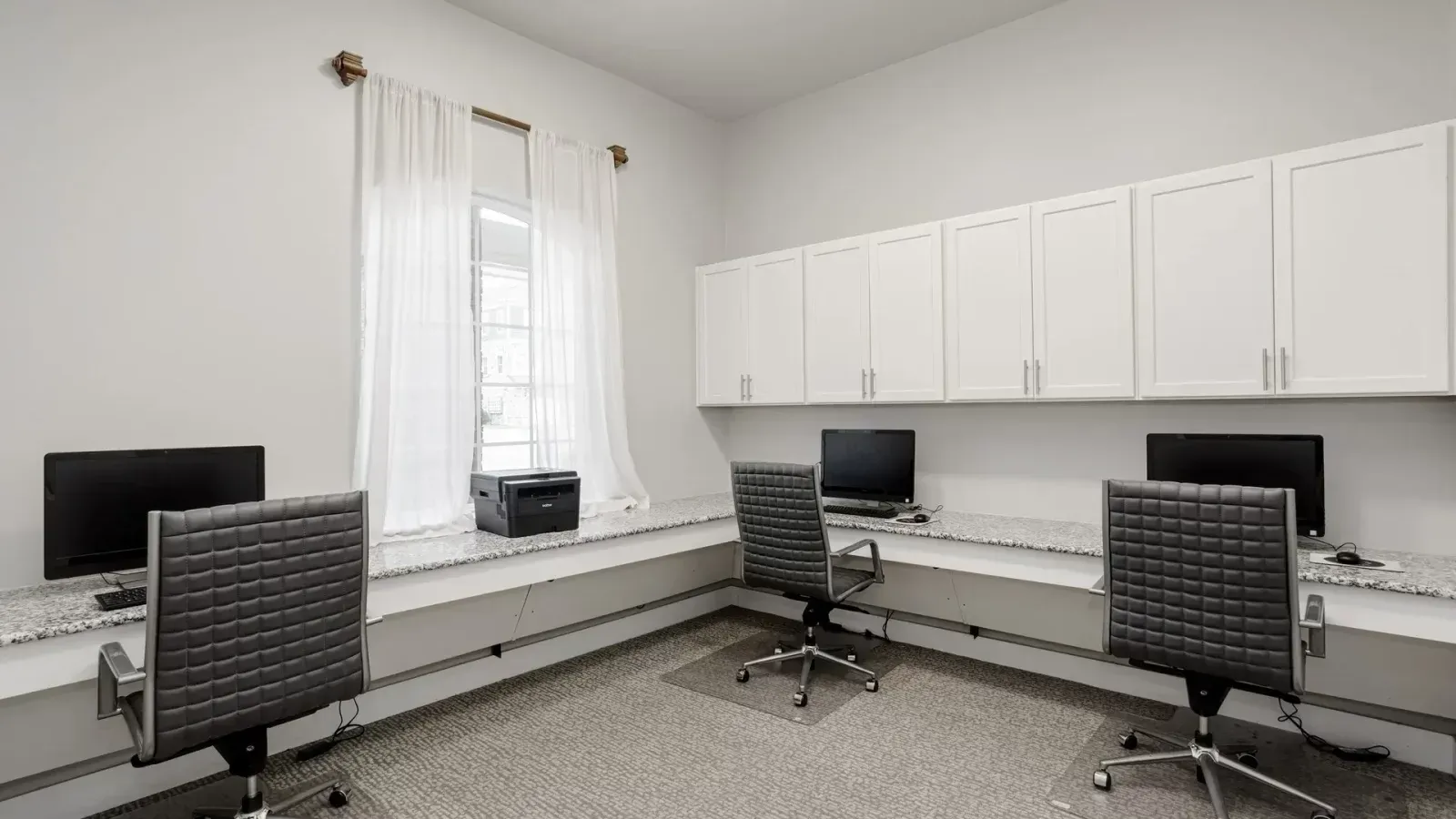 Bright office workspace with three desks, computers, rolling chairs, and white cabinets along the wall