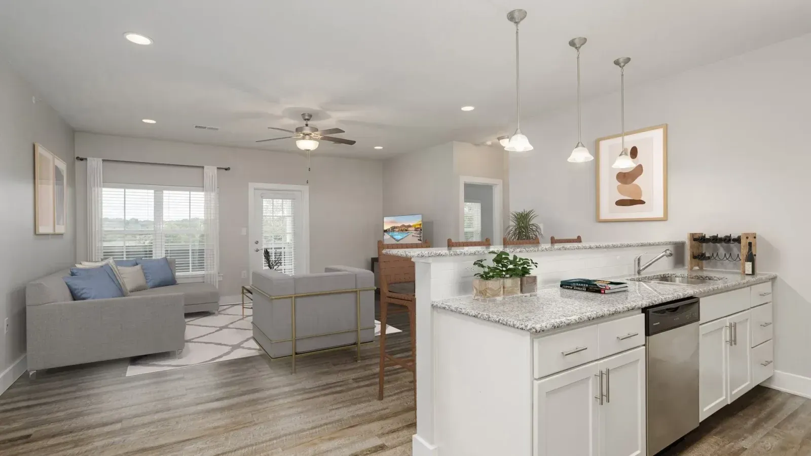 Bright open-concept kitchen and living room with white cabinets, gray sofas, wood floors, and pendant lights